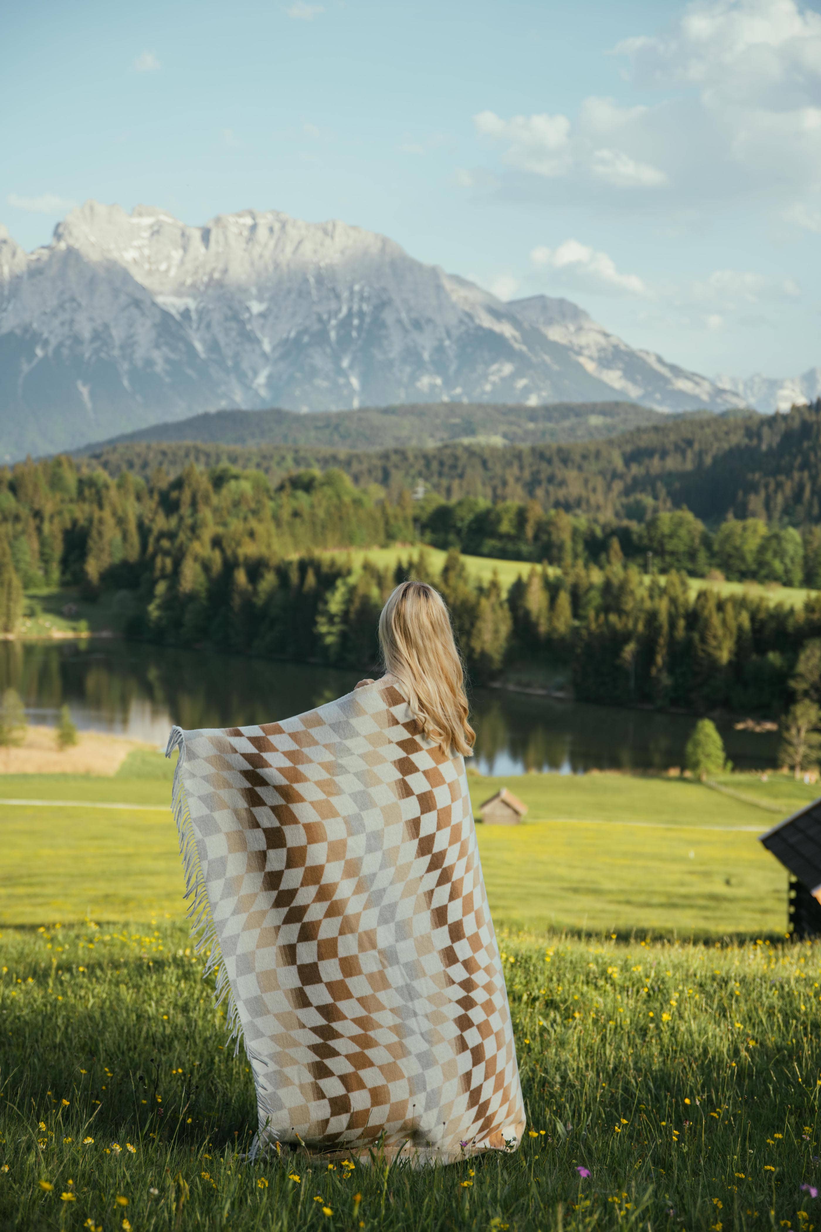 A woman holds a Checkered Palm Desert blanket in a field, with a lake and mountains in the background. Sustainable materials, 72”l x 60”w size, and recycled wool blend.
