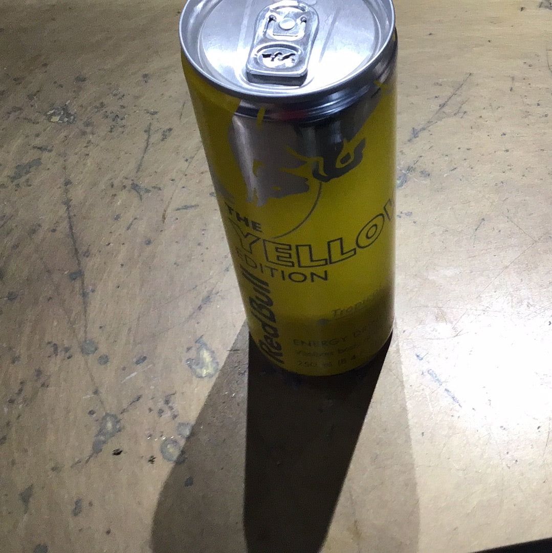 A close-up of a Red Bull Tropical 8.4oz aluminum can on a table, featuring black text, a yellow surface, and a beverage can.