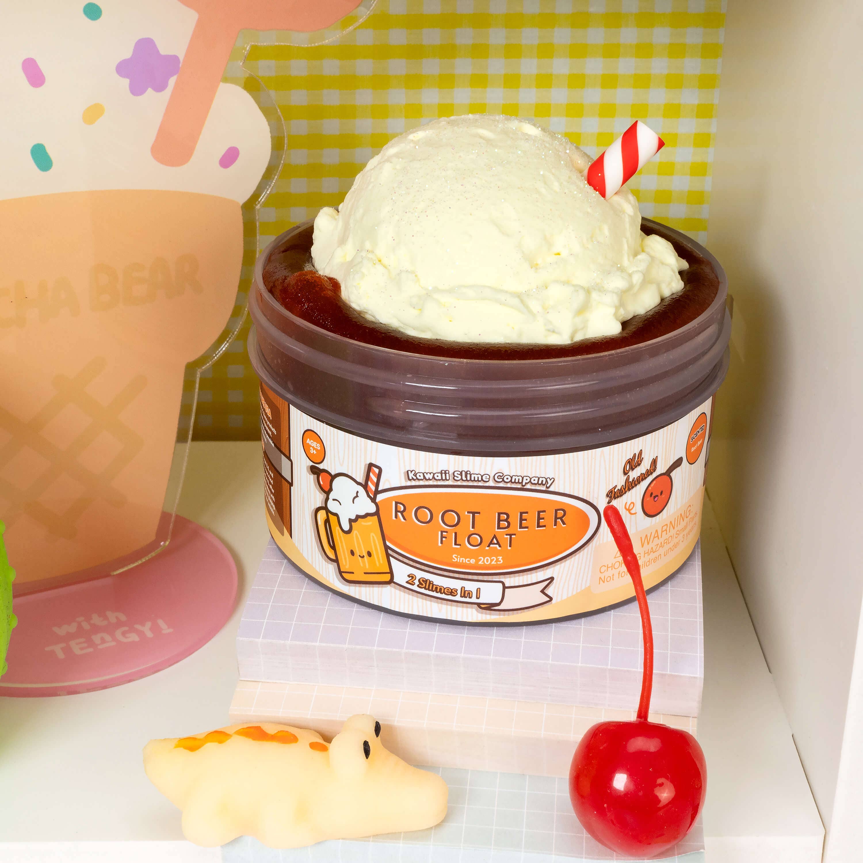 A glossy slime container with a cherry and toy, accompanied by a straw, reflecting a bear on a mirror. Product: Root Beer Float 2 Slimes In 1.