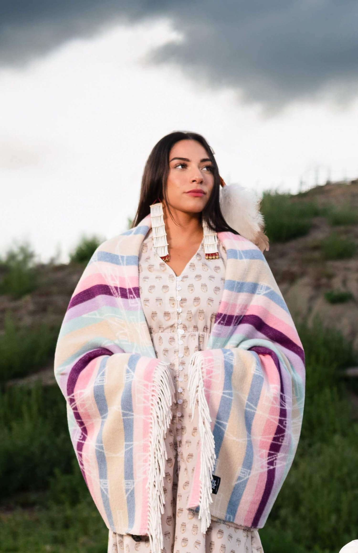 A woman in a dress with a colorful blanket by Lauren Good Day, featuring Hunaaneeká (Spring) design. Made from recycled materials in a sustainable process. Size: 72”l x 60”w.