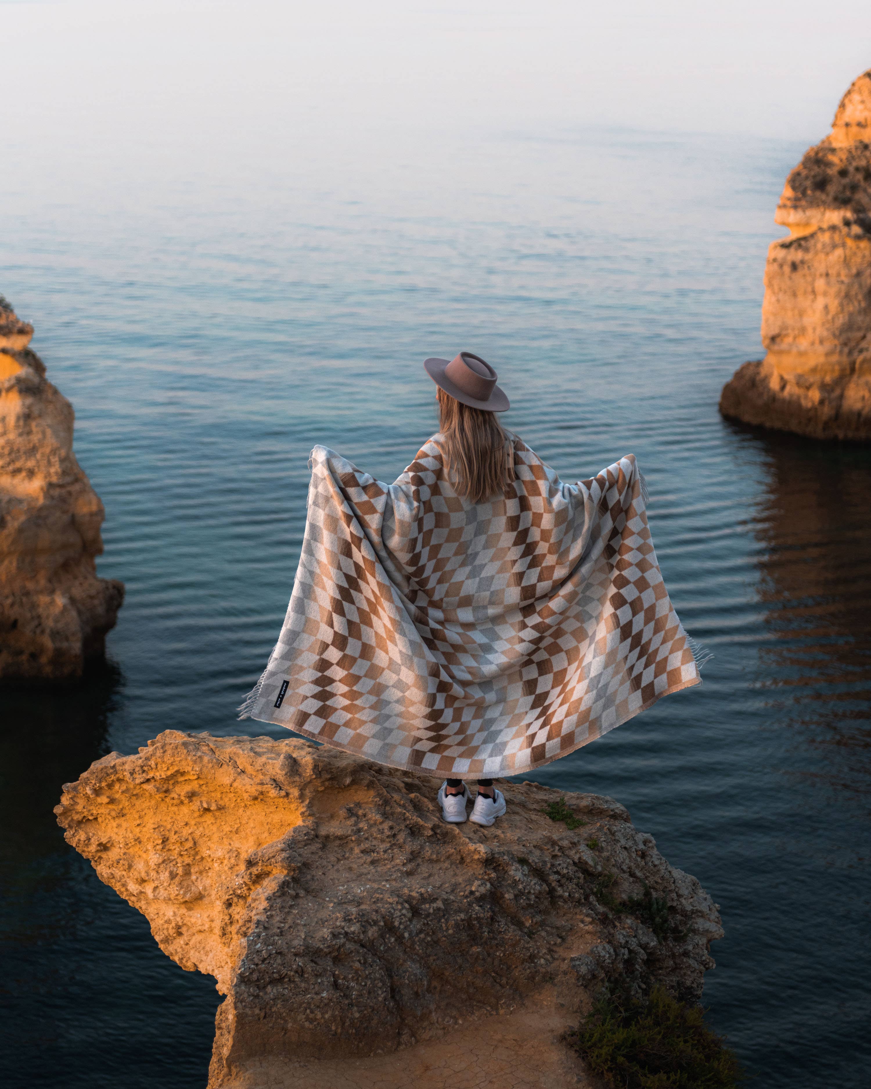 A woman sits on a rock overlooking water, draped in a Checkered Palm Desert blanket. Sustainable materials, 72”l x 60”w size, and a commitment to donate blankets to shelters.