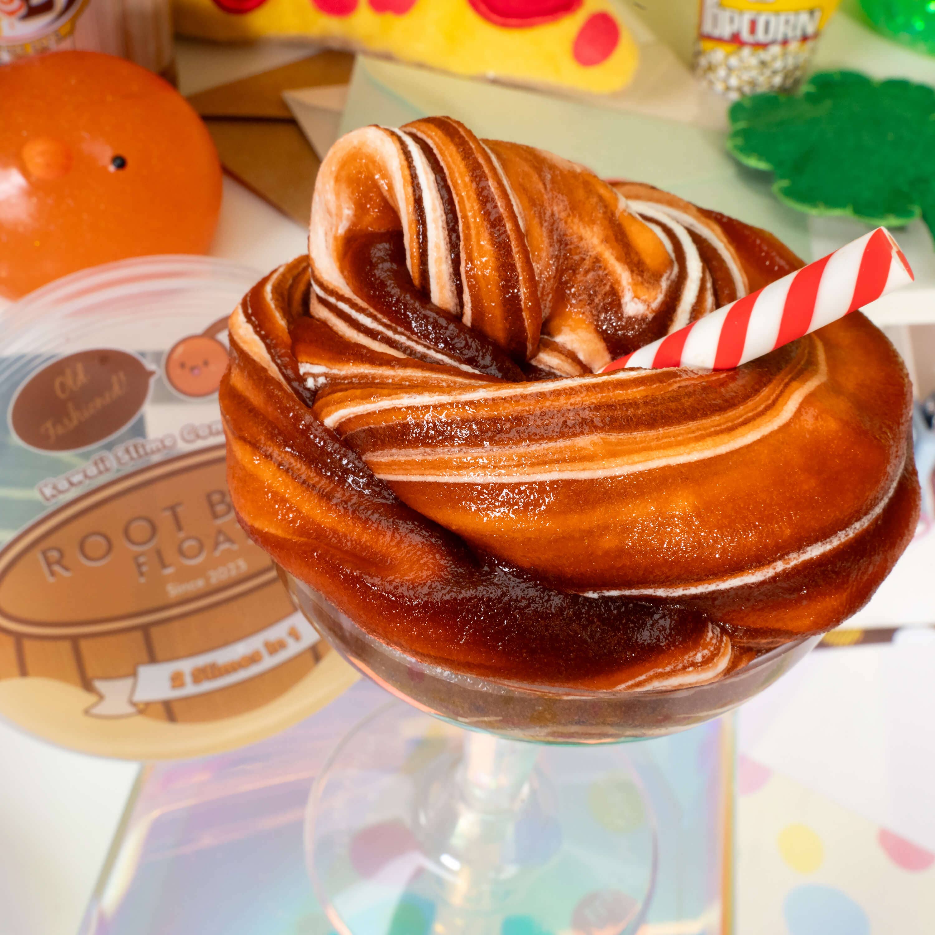 A close-up of a brown and white swirly slime in a glass jar, accompanied by an ice cream scoop clay topper. Root Beer Float 2 Slimes In 1 product.