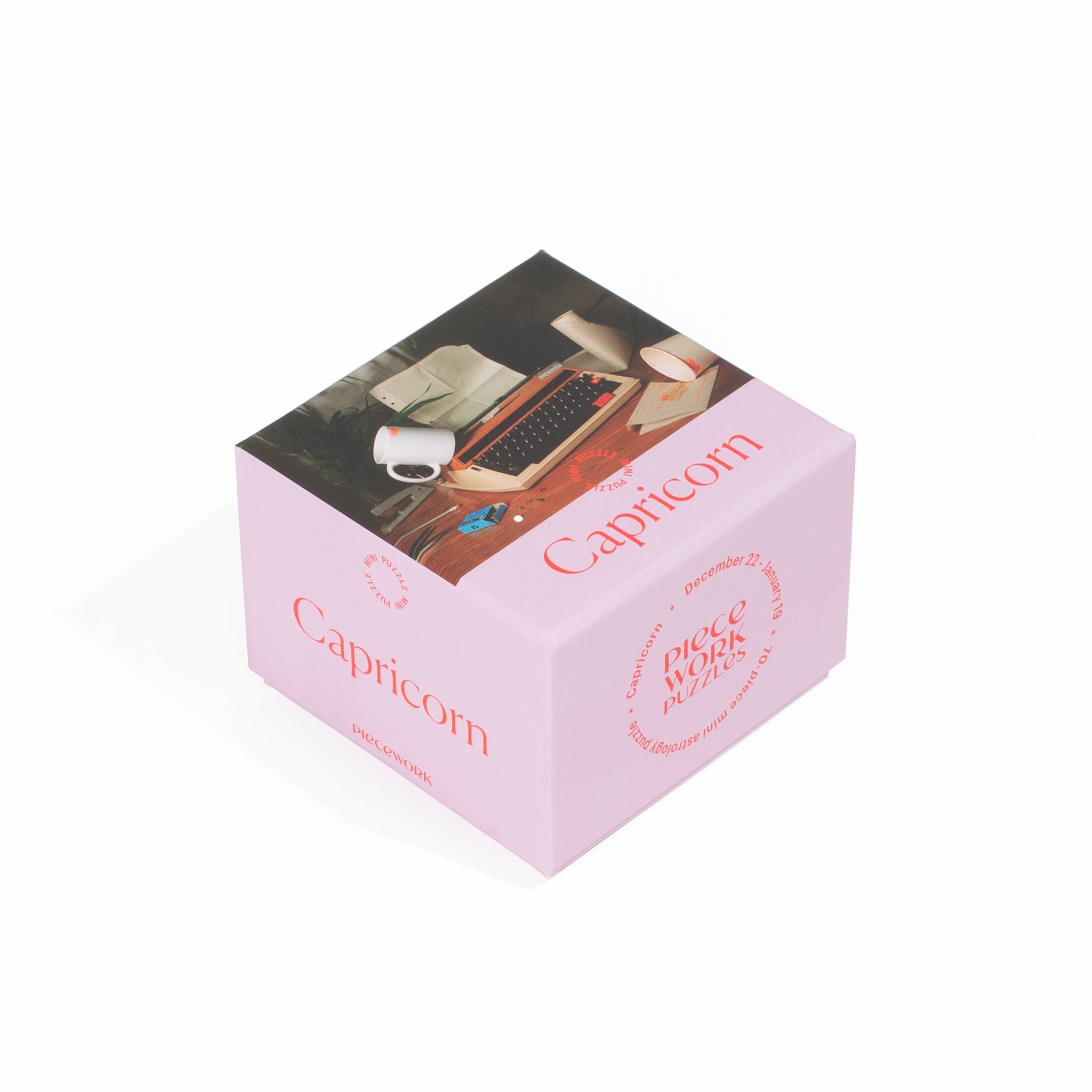 A pink box with a Capricorn Mini Puzzle featuring a typewriter and a mug. Puzzle size 5 x 7 inches, box dimensions 3.5 x 2.5 x 2.75 inches, recycled paper board, 70 pieces.