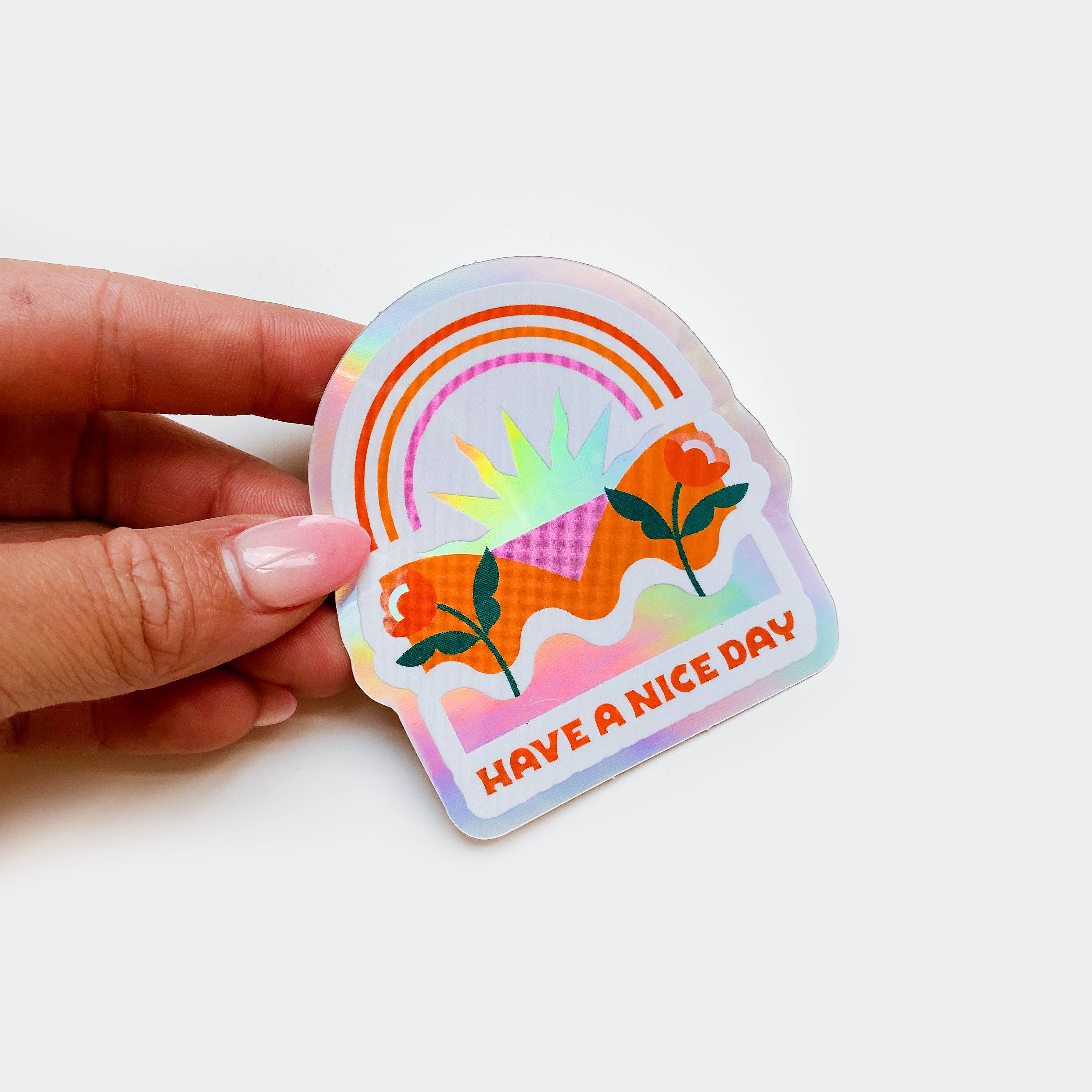 A hand holds a die-cut holographic sticker featuring a sunrise design. Glossy coated vinyl, waterproof, 2.5 x 2.75 size.