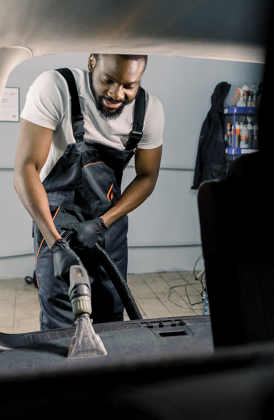 A man in overalls uses a vacuum cleaner to deep clean a car carpet. Service includes steam extraction and stain removal. Approximate time: 30 minutes - 1 hour.
