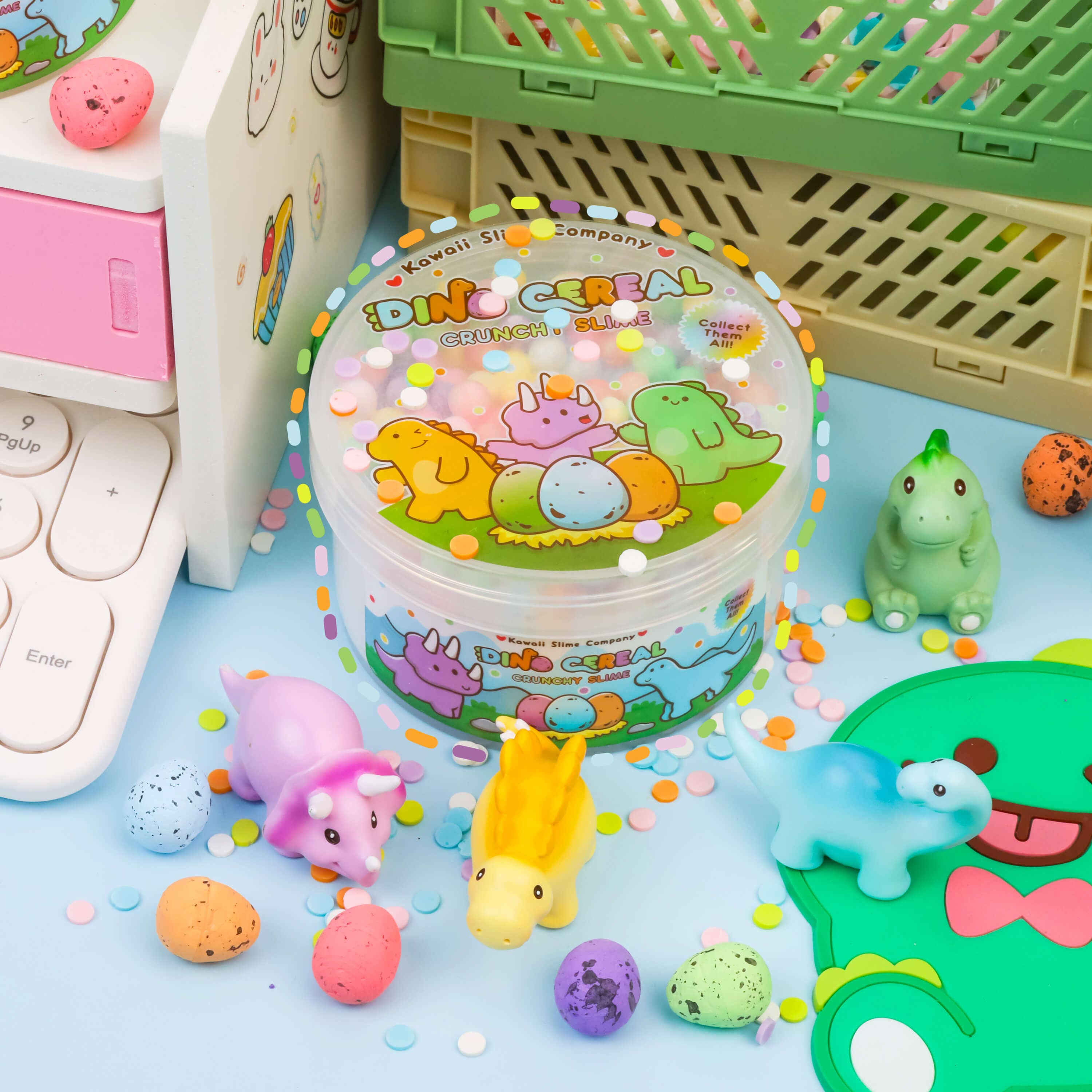 A group of baby toys including green, pink, and yellow dinosaur figures, a white egg, and a purple ball, displayed next to a keyboard. Product: Dino Cereal Crunchy Slime with collectible dinosaur miniatures and confetti sprinkles.
