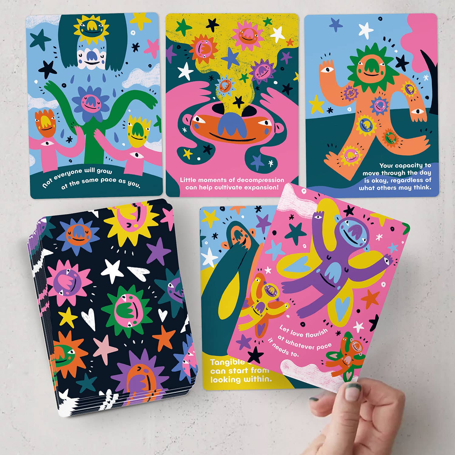 A hand holding the Gentle Reminders Deck by Barry Lee, a deck of emotional self-care cards with rounded corners, featuring illustrations of stars, sun, and cartoon characters.