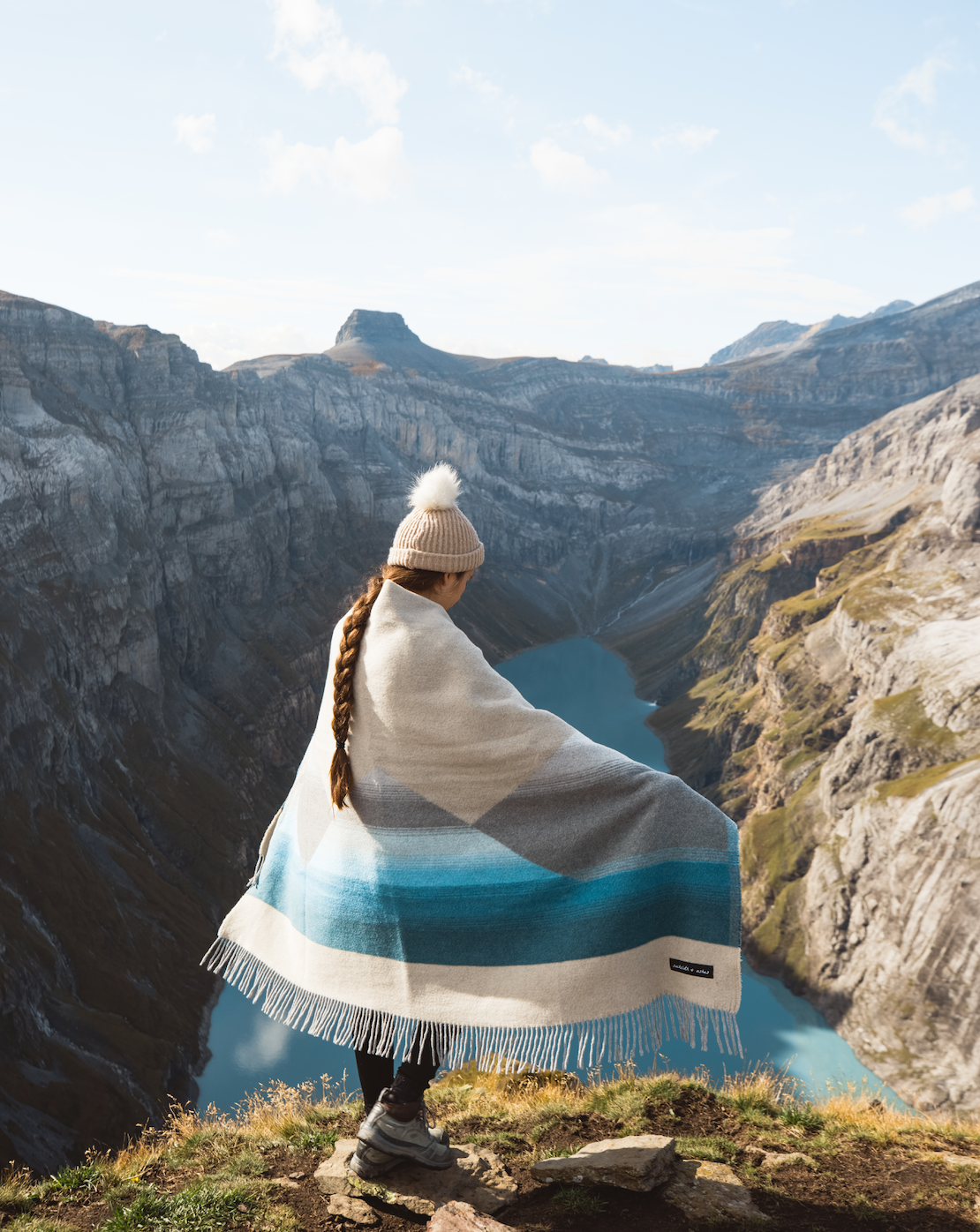 A woman stands on a cliff overlooking a river, embodying the spirit of our Mountain Arctic blanket. Recycled wool blend, 70”l x 60”w, for adventures and giving back.