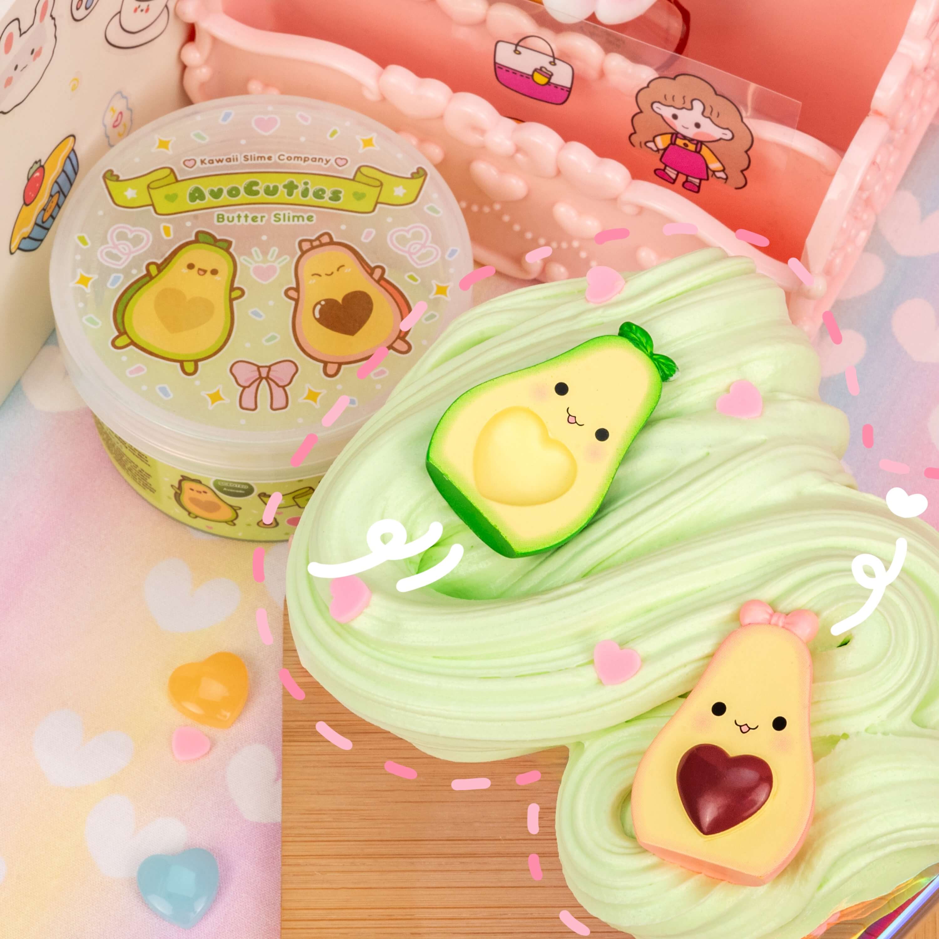 AvoCuties Butter Slime (4pcs/case) featuring an avocado-shaped toy, cartoon characters on a plastic container, and a heart-shaped charm on fabric. Creamy, fluffy, and scented for a delightful play experience.