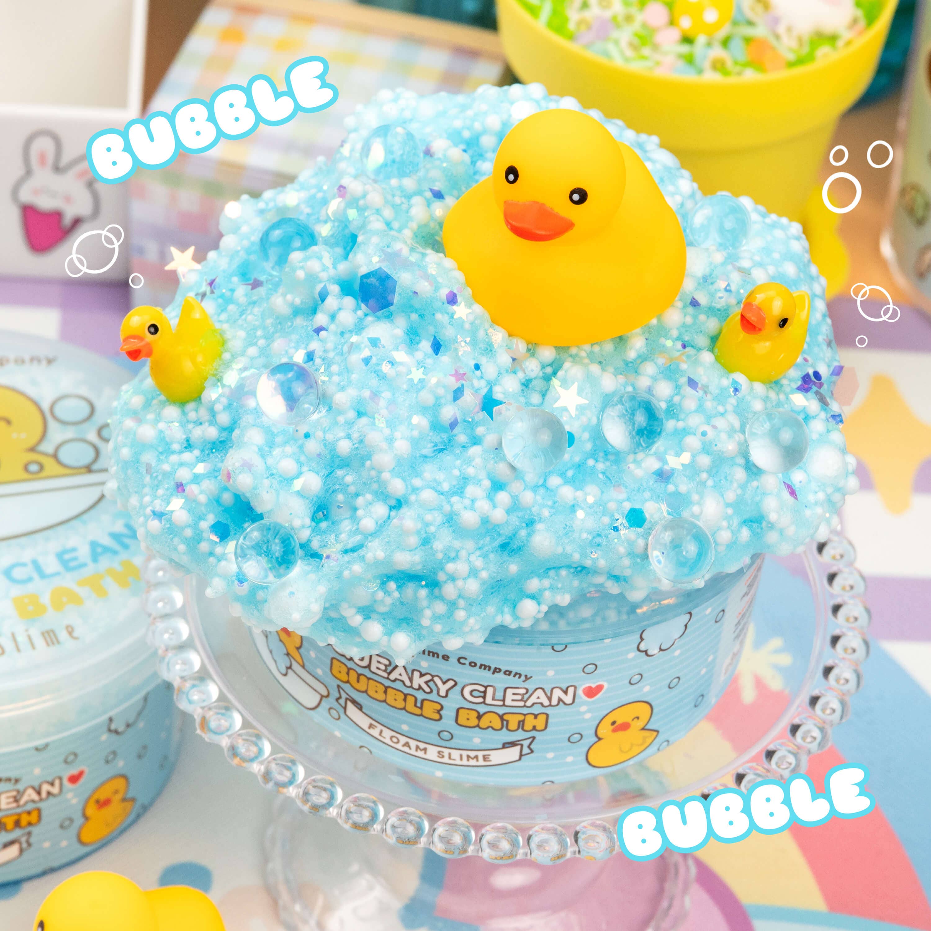 A blue bubble bath with a yellow rubber duck on top, part of Squeaky Clean Bubble Bath Floam Slime. Includes foam beads, glitter, and a floral scent. Cute squeaky duck toy included.