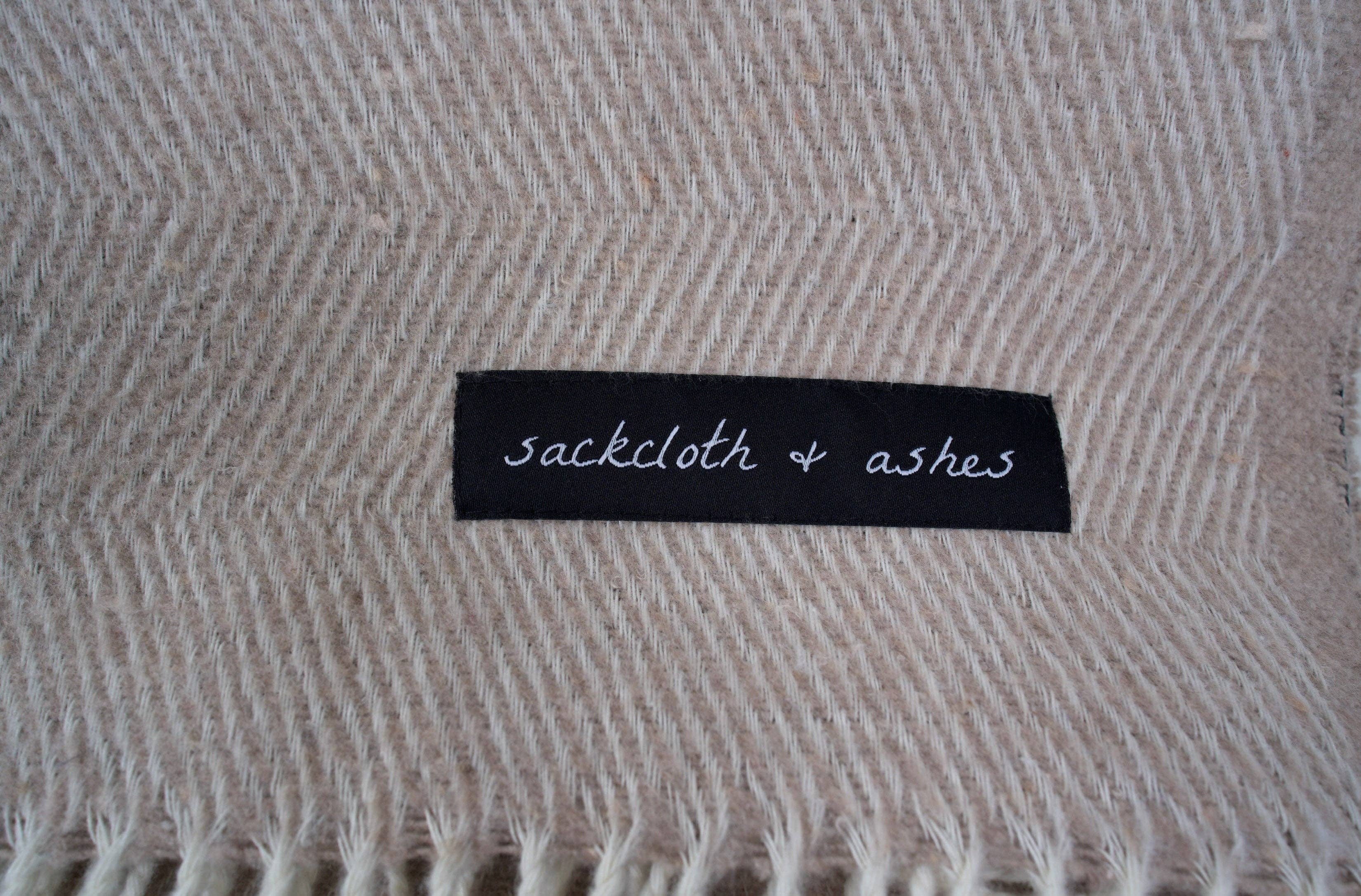 Close-up of Herringbone Sand blanket label on black fabric, featuring elegant white text. Made in Italy from recycled cotton and polyester, water-free process, emphasizing comfort and durability.