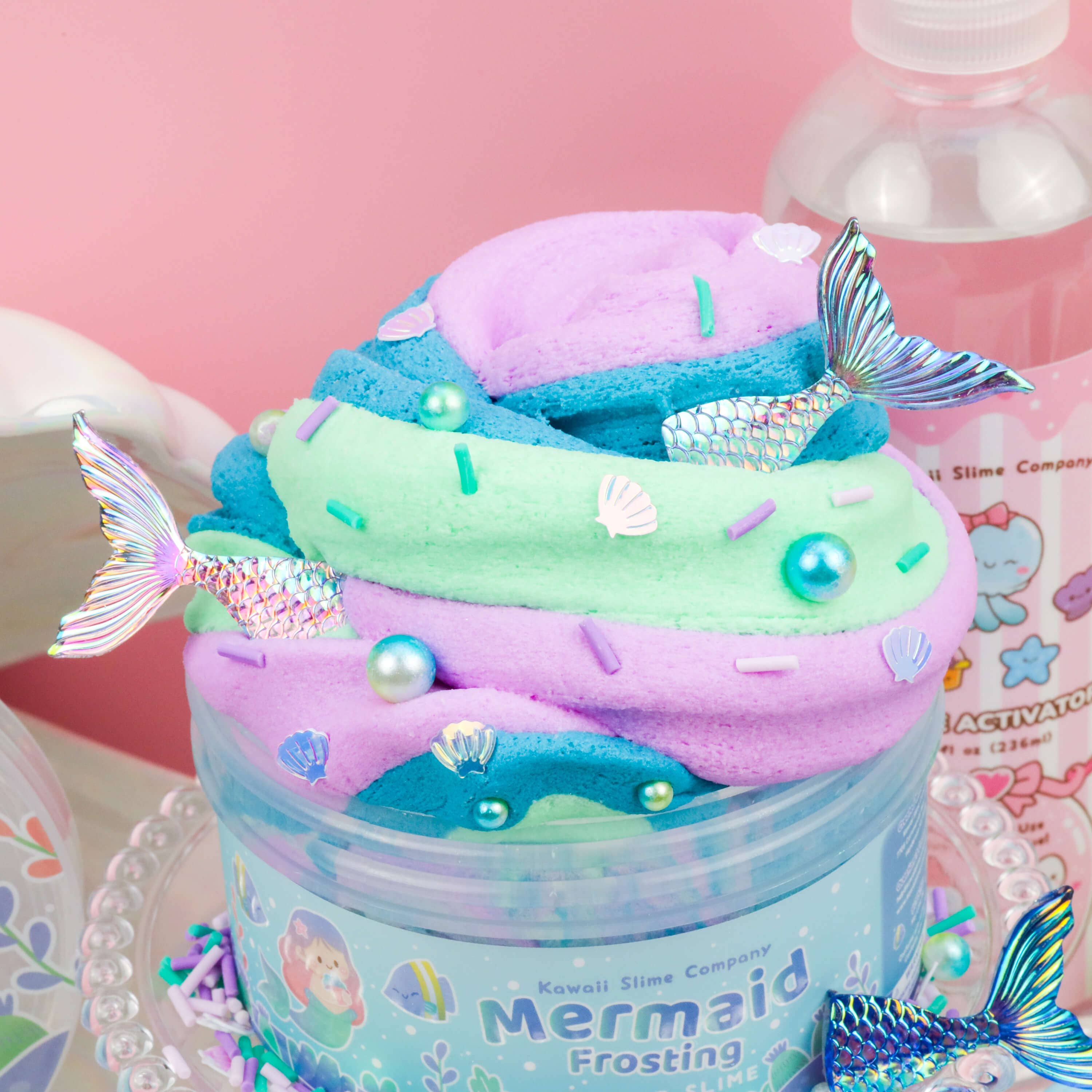 A jar of colorful "Mermaid Frosting Butter Slime (4pcs/case)" with blue, pink, and green swirls, decorated with small silver mermaid tail charms, pearls, and magical mermaid toppings. The jar is labeled "Mermaid Frosting" from Kawaii Slime Company and nearby is a bottle of "Slime Activator." The background is light pink.