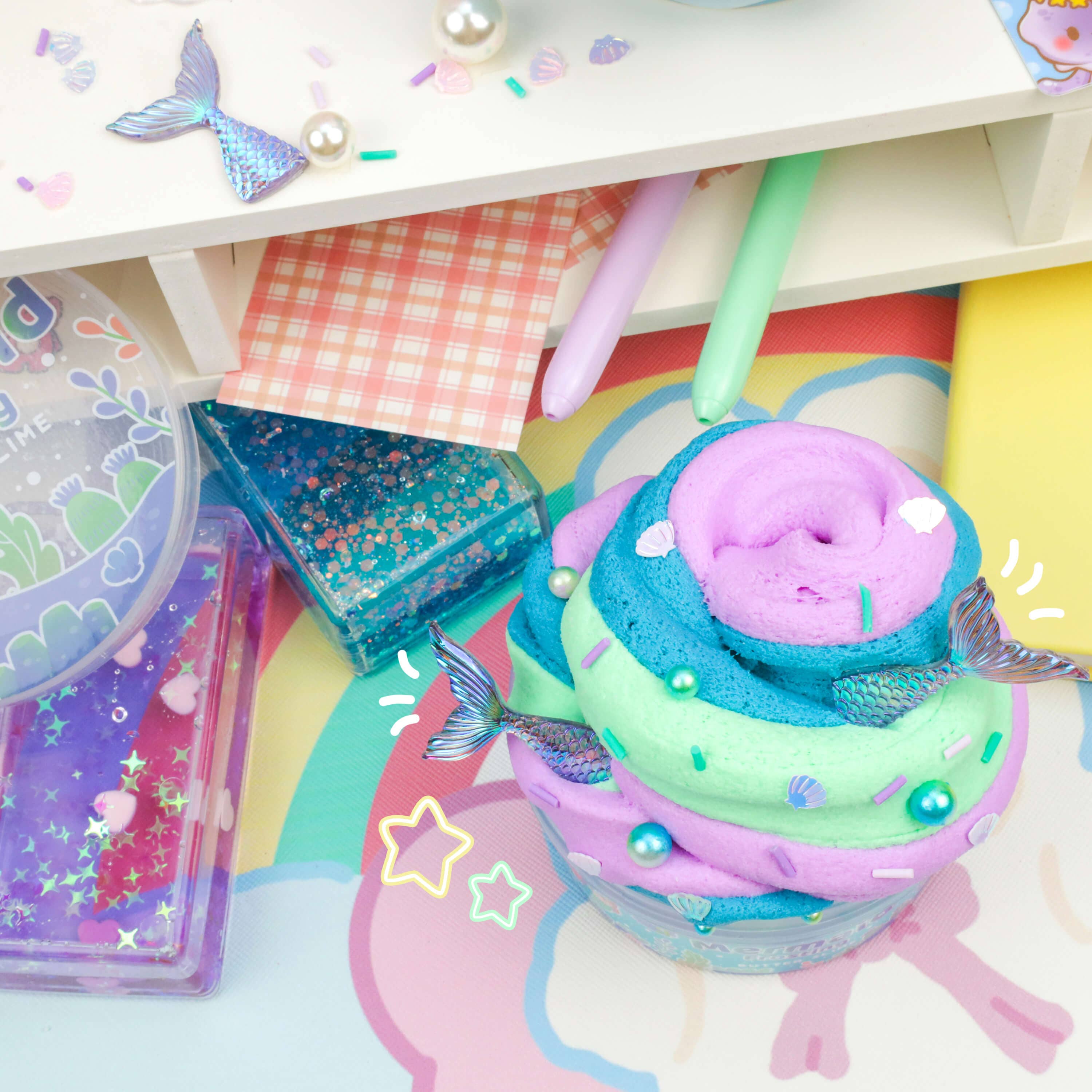 An image of a colorful, swirl-shaped slime in pastel shades of blue, green, and purple. This Mermaid Frosting Butter Slime (4pcs/case) from Kawaii Slime Company is adorned with magical mermaid toppings like tiny tails, sprinkles, and decorative pearls. Surrounding it are various items such as glittery pens, checkered paper, and containers of glitter.