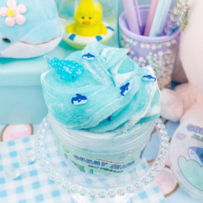 A blue and white slime filled with shark fimo slices and whale shark charms, creating a mesmerizing ocean swirl. Jelly Creme texture with meringue pieces, scented like blue raspberry candy and marshmallow cream.