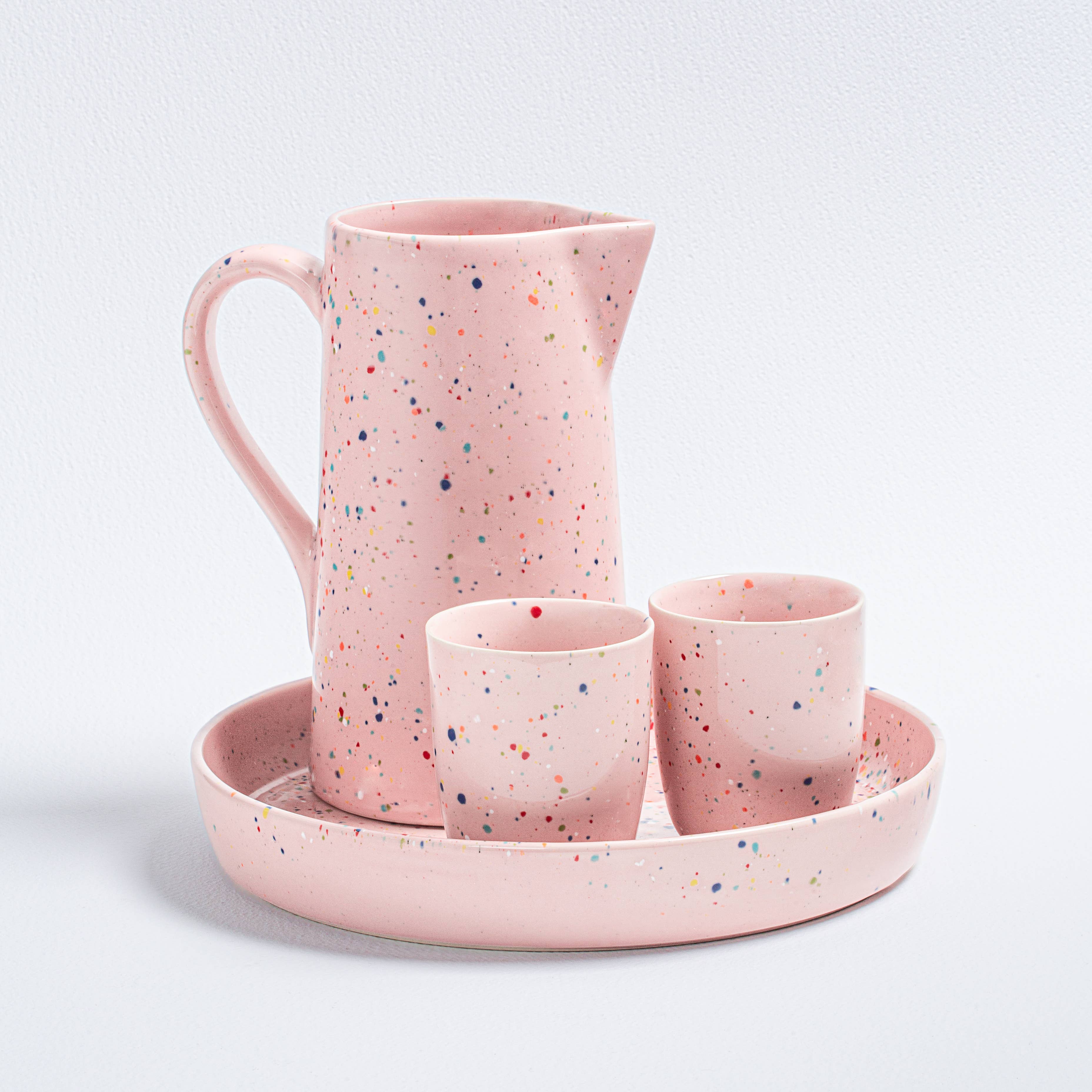 A pink earthenware pitcher and cup set on a plate, showcasing speckled design. Ideal for everyday use, dishwasher and microwave safe. Capacity: 1.5L. Celebrate uniqueness with artisanal touch.