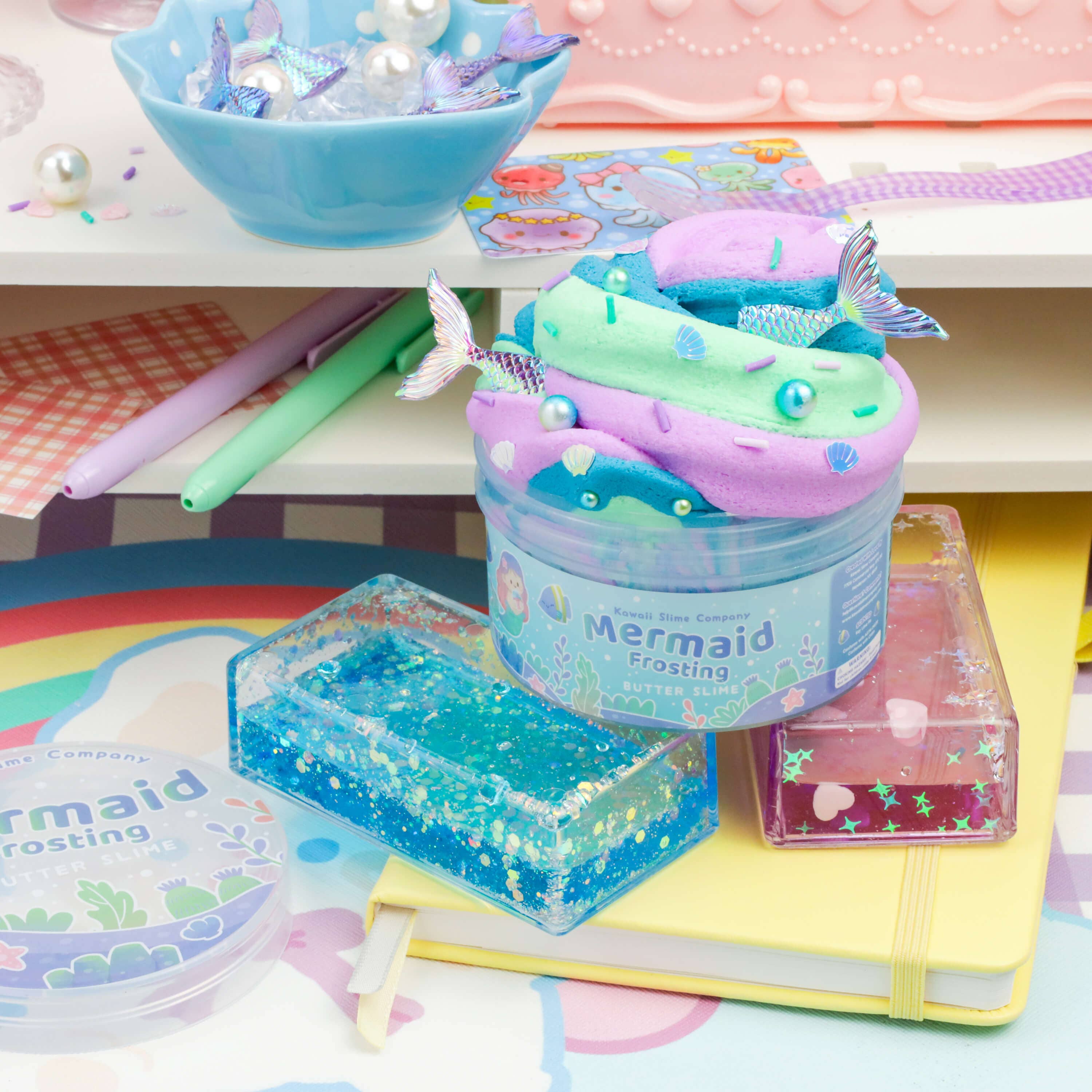 A colorful desk scene features a jar of "Mermaid Frosting Butter Slime (4pcs/case)" by Kawaii Slime Company. The teal and lavender concoction with mermaid tails resembles magical mermaid toppings. Nearby, pens, a notebook, holographic clear blue and pink containers, and a bowl with more slime and seashells complete the enchanting setup.