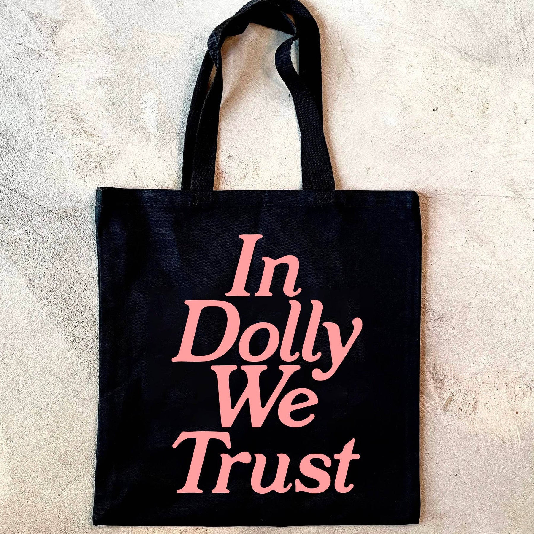 A black tote bag with pink text In Dolly We Trust and a woven shoulder strap. Durable and spacious for all your essentials.