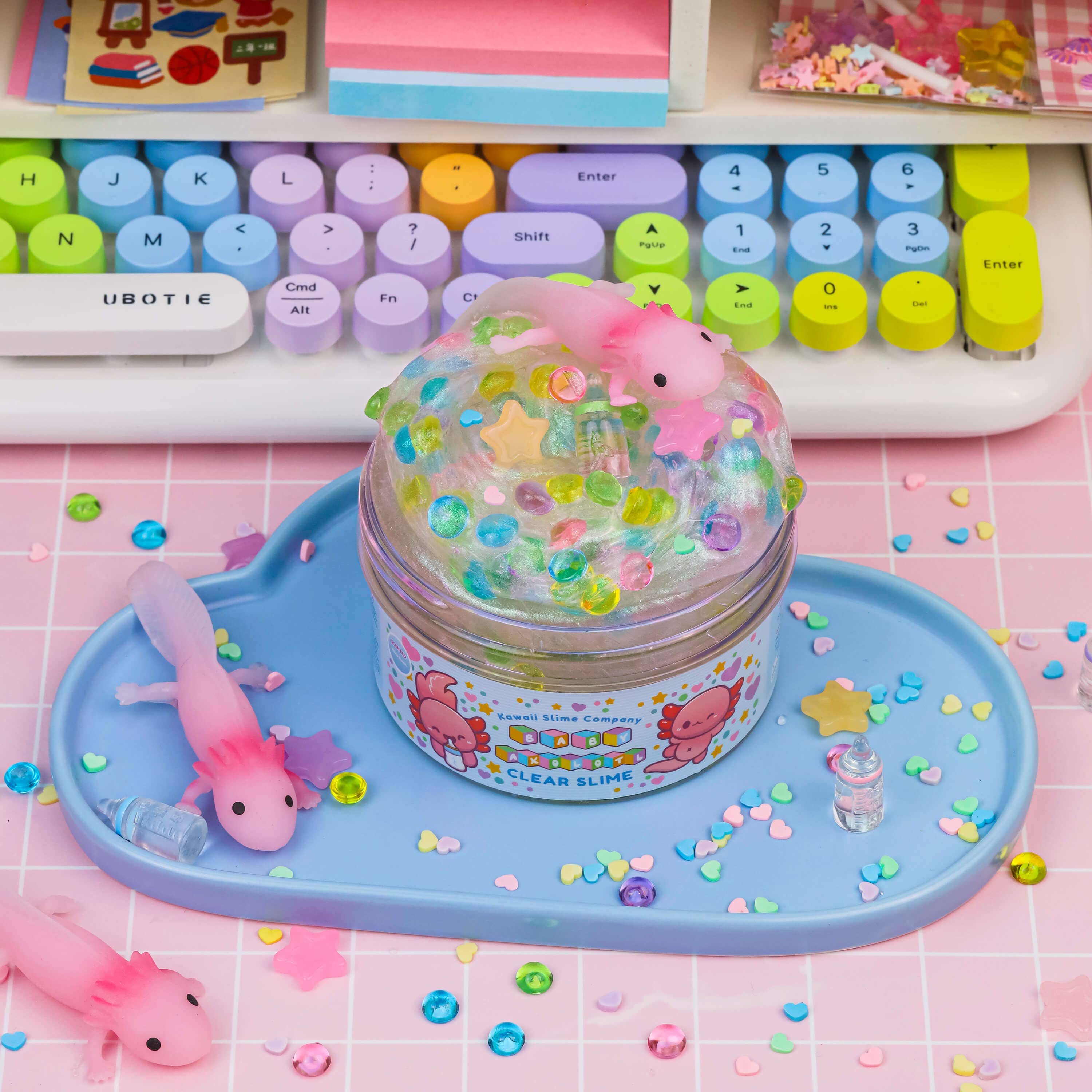 A jar of clear slime with small pink toy animals on top, resembling baby axolotls, set on a blue tray. Cute and easy-to-care-for underwater pet-themed slime.
