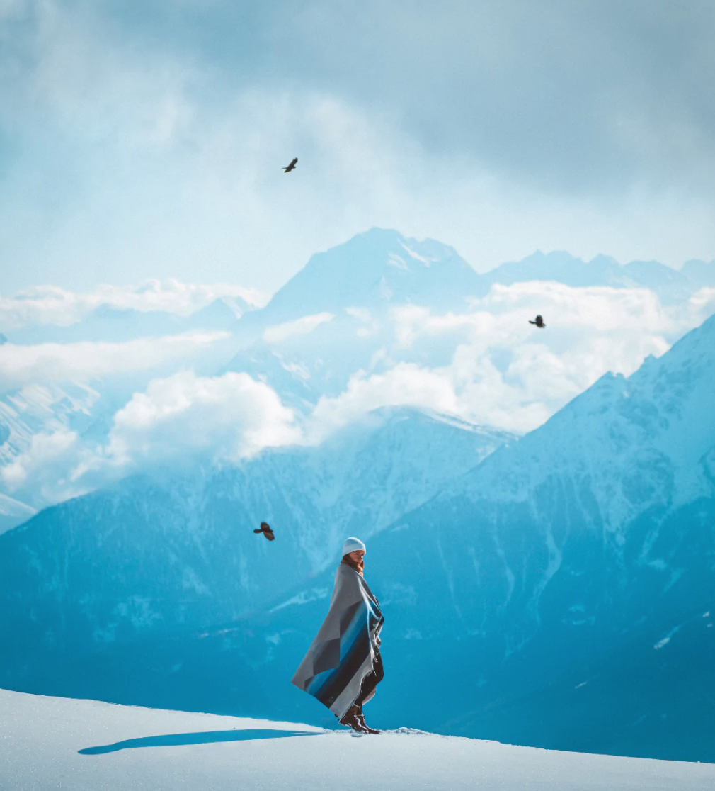 A person on snowy mountain with birds, a flag waving; Mountain Arctic blanket made from recycled materials, 70”l x 60”w, 3 lbs, Recycled Wool/Polyester/Acrylic.