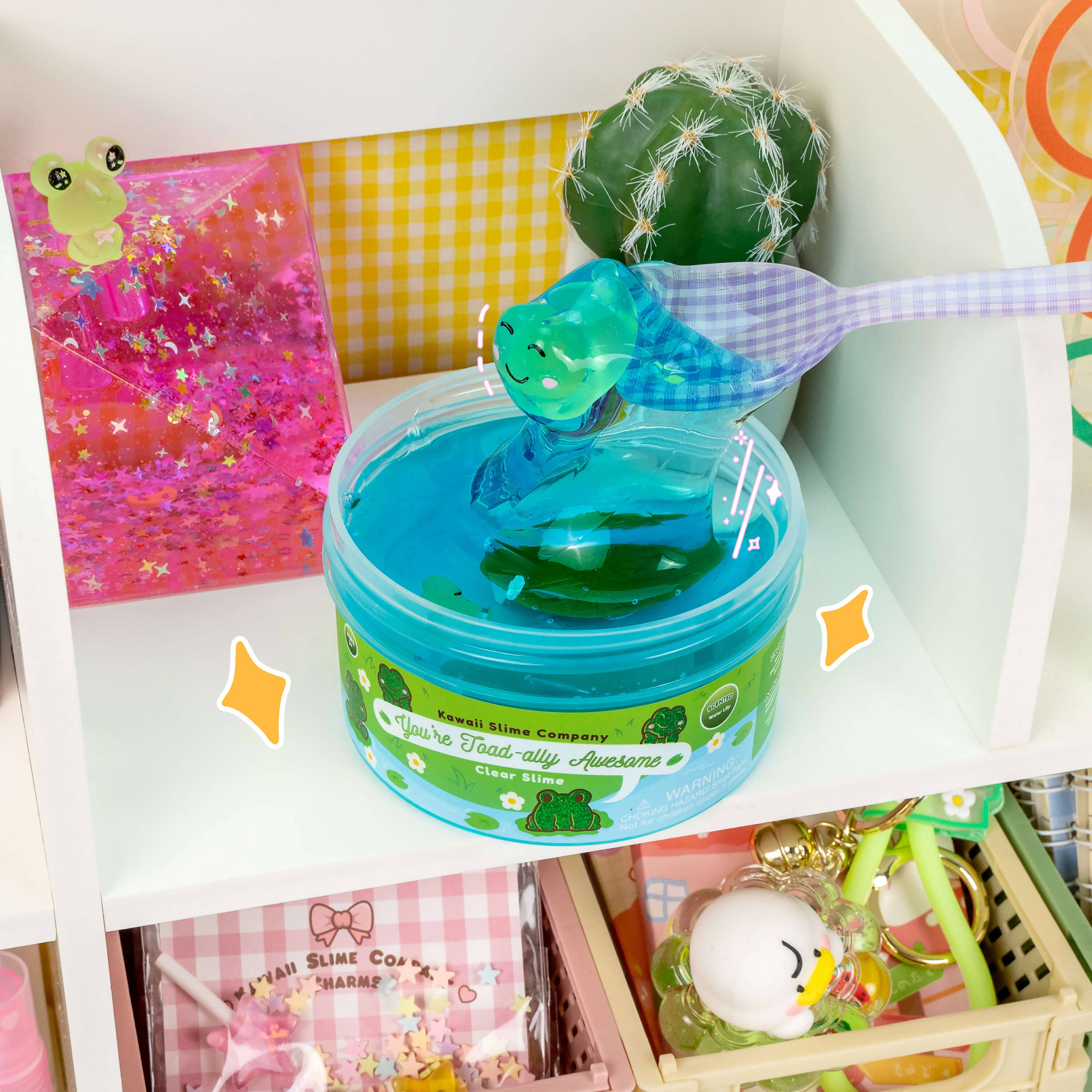 A clear plastic container with blue slime, two toad charms, and a spoon inside. Textured for poking and popping bubbles, scented like water lilies for a calming playtime experience.