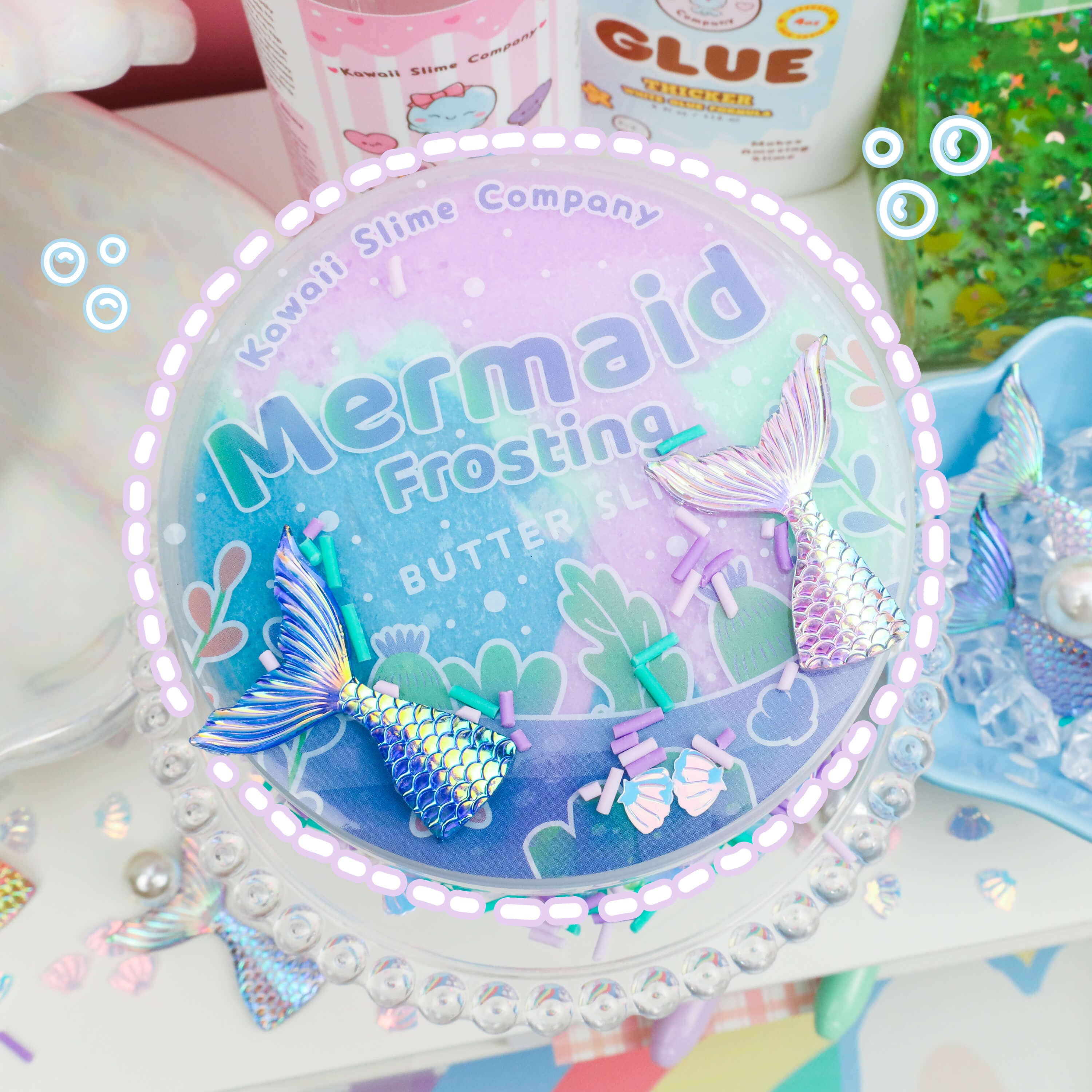 A container of "Mermaid Frosting Butter Slime (4pcs/case)" from Kawaii Slime Company is surrounded by glittering decorations and slime supplies, including glue and floating beads. The lid features a gradient design with mermaid tails and sprinkles, complemented by magical mermaid toppings for an enchanting touch.