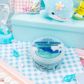 A Gummy Shark Jelly Creme Slime featuring shark fimo slices in a vibrant ocean-themed slime with whale shark charms. Cool, swirled texture scented like blue raspberry candy and marshmallow cream.