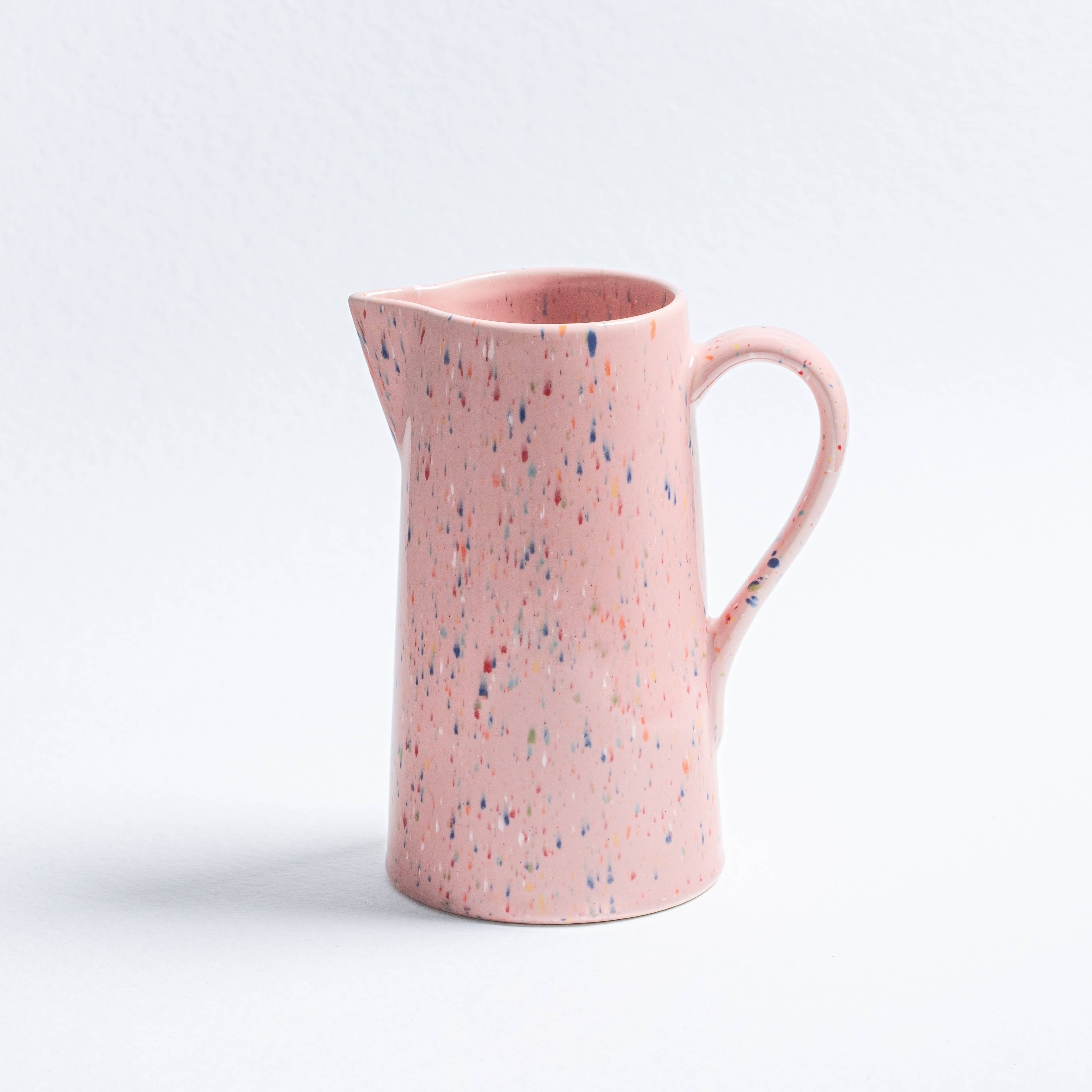 A pink earthenware pitcher with multicolored speckled spots and a handle, perfect for everyday use. Dishwasher and microwave safe, 1.5L capacity. Celebrate individuality with unique artisanal design.