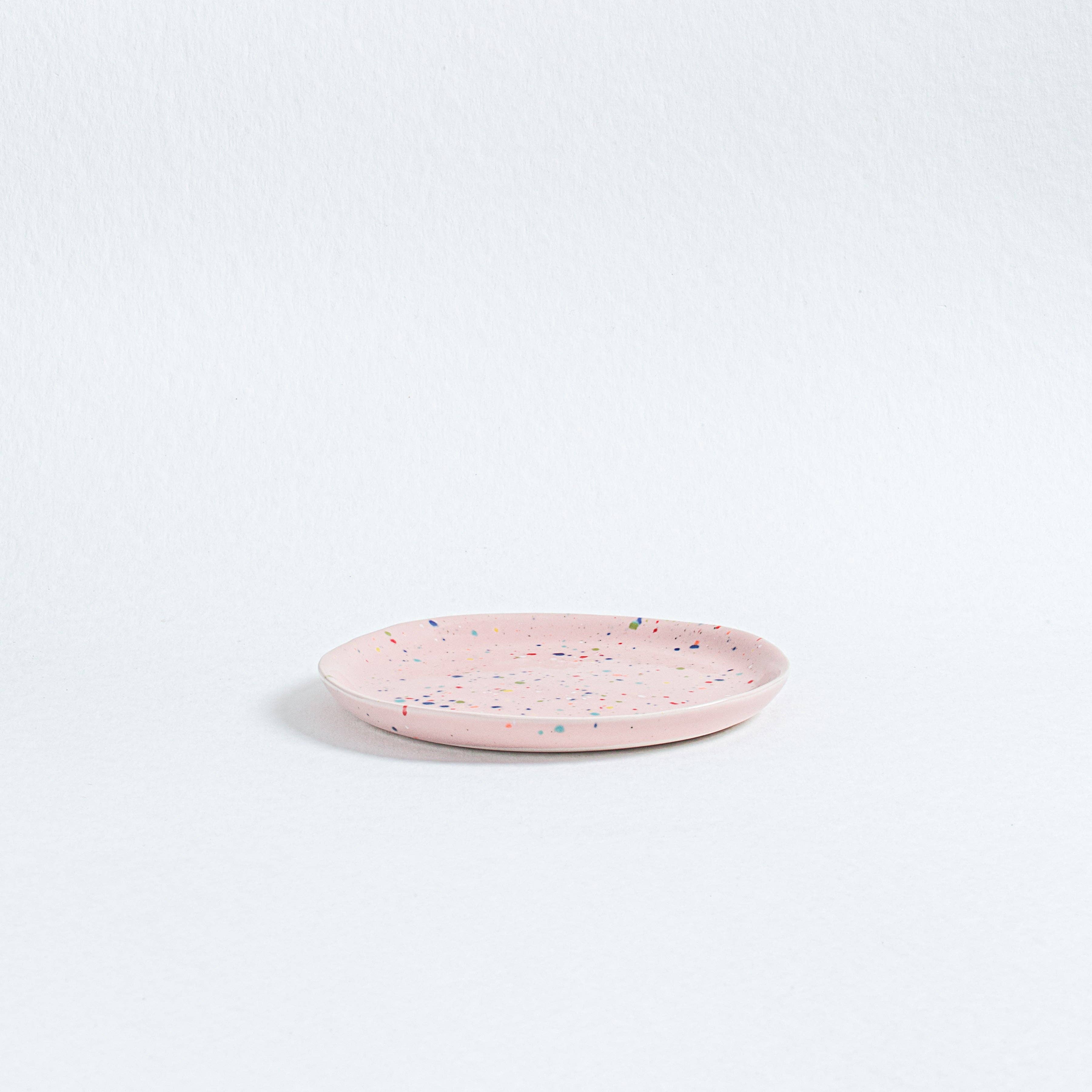 A pink ceramic bread plate with multicolored speckled spots, part of the vibrant Party collection. Diameter: 17cm, Height: 1.6cm. Dishwasher and microwave safe, food safe, and Cadmium Free. Crafted uniquely, each plate varies slightly.