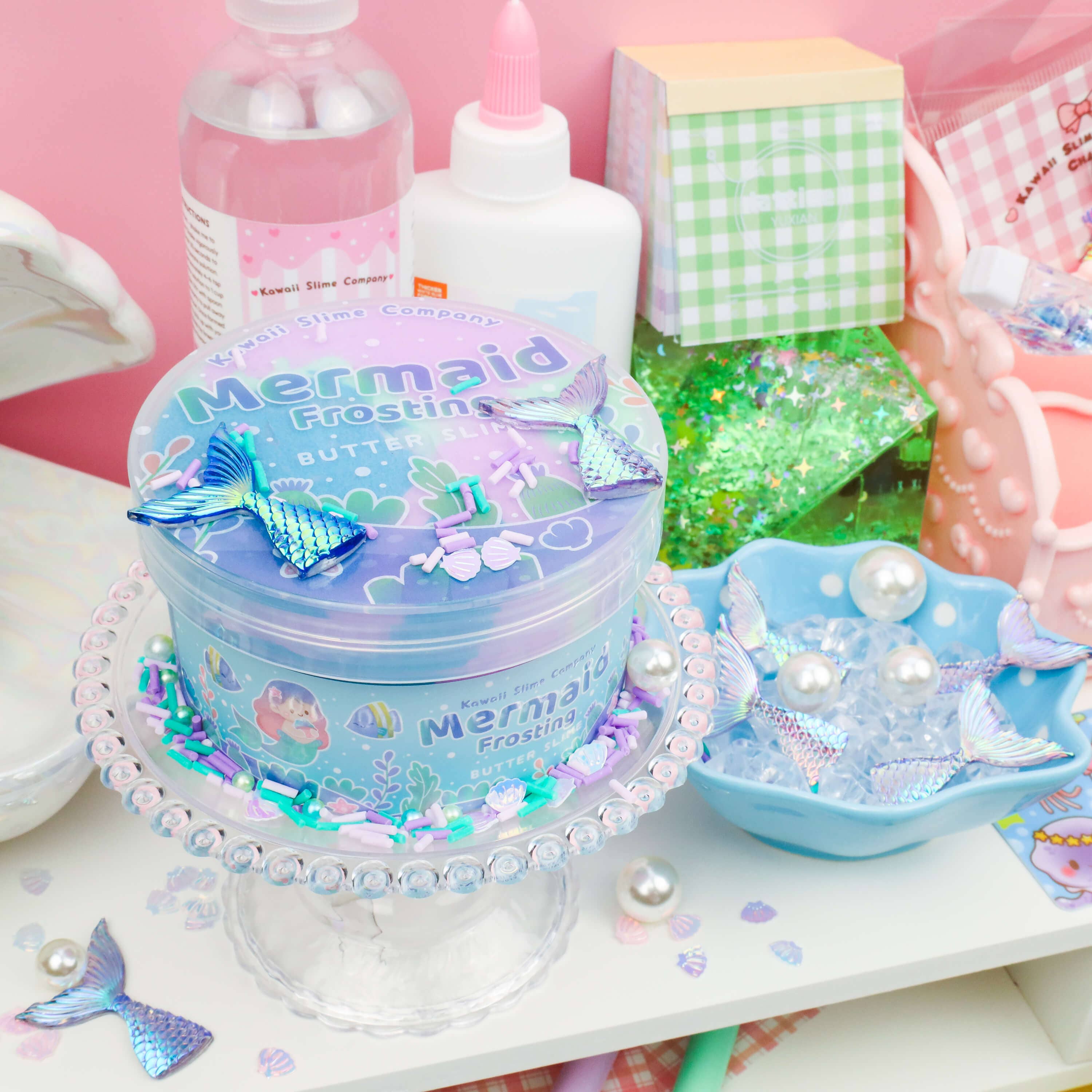 A pastel-themed craft workspace features a container labeled "Mermaid Frosting Butter Slime (4pcs/case)" from Kawaii Slime Company on a decorative stand. The table is adorned with mermaid tail charms, iridescent beads, and craft supplies, including glue, glitter paper, and a spray bottle, set against a pink wall.