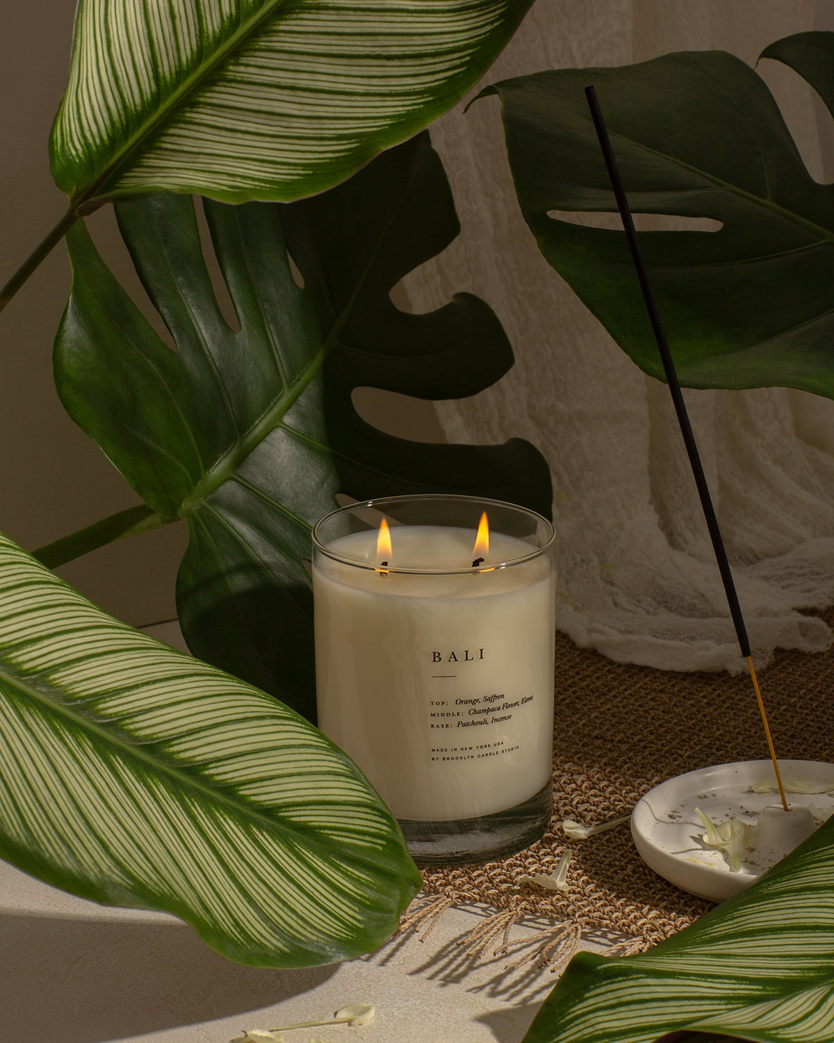 A serene scene featuring a candle, leaf, and flower. Bali Escapist Candle: Orange, saffron, champaca flower, incense notes. Handcrafted in Brooklyn with soy wax. Vegan and eco-friendly.