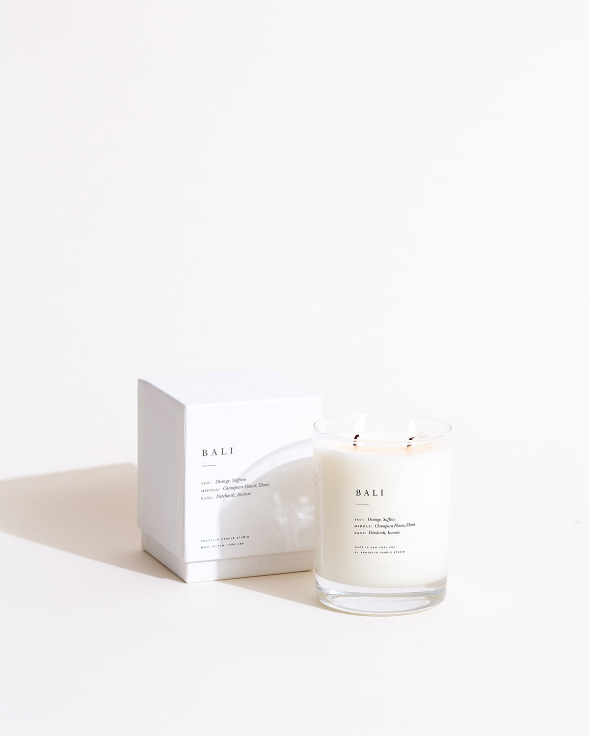 A white candle in a glass container, embodying the Bali Escapist Candle's blend of orange, saffron, champaca flower, elemi, patchouli, and incense notes.