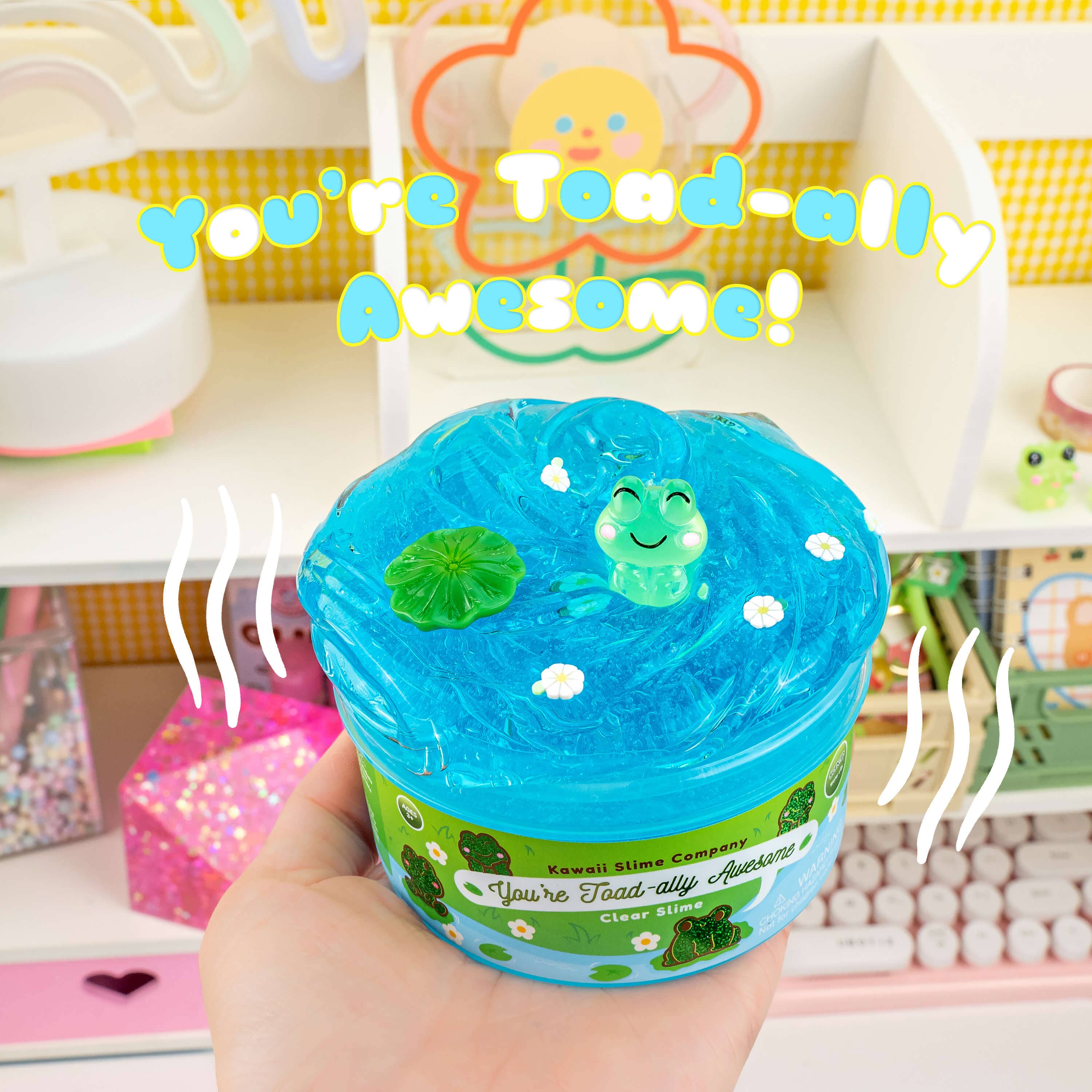 A hand holding a jar of You're Toad-ally Awesome Clear Slime with two toad charms, featuring a refreshing water lily scent and bright blue color.