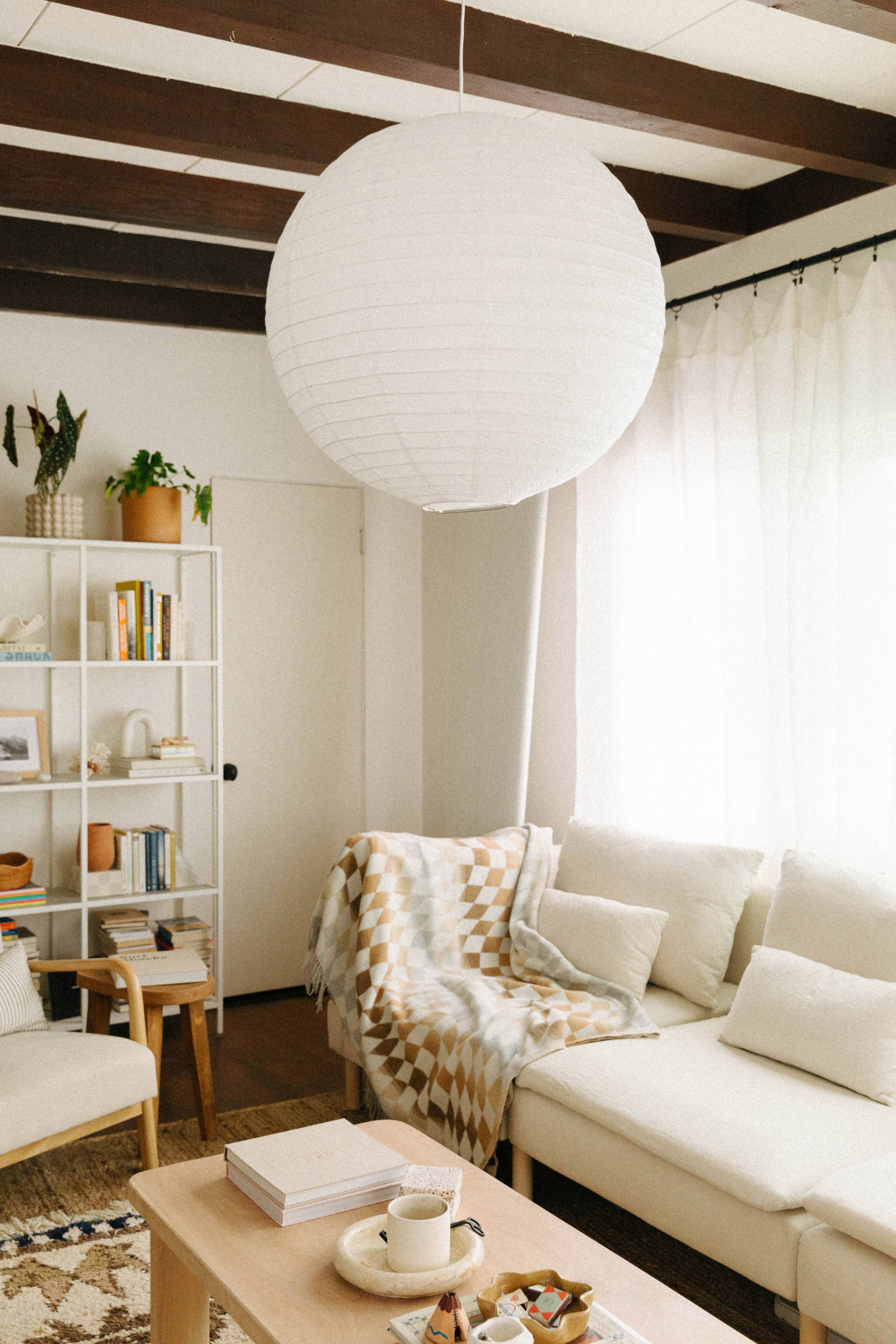 A white round paper lantern hangs from a ceiling in a living room, surrounded by a cup on a saucer and a stack of white paper. Size: 72”l x 60”w. Contents: Recycled Wool / Polyester / Acrylic.