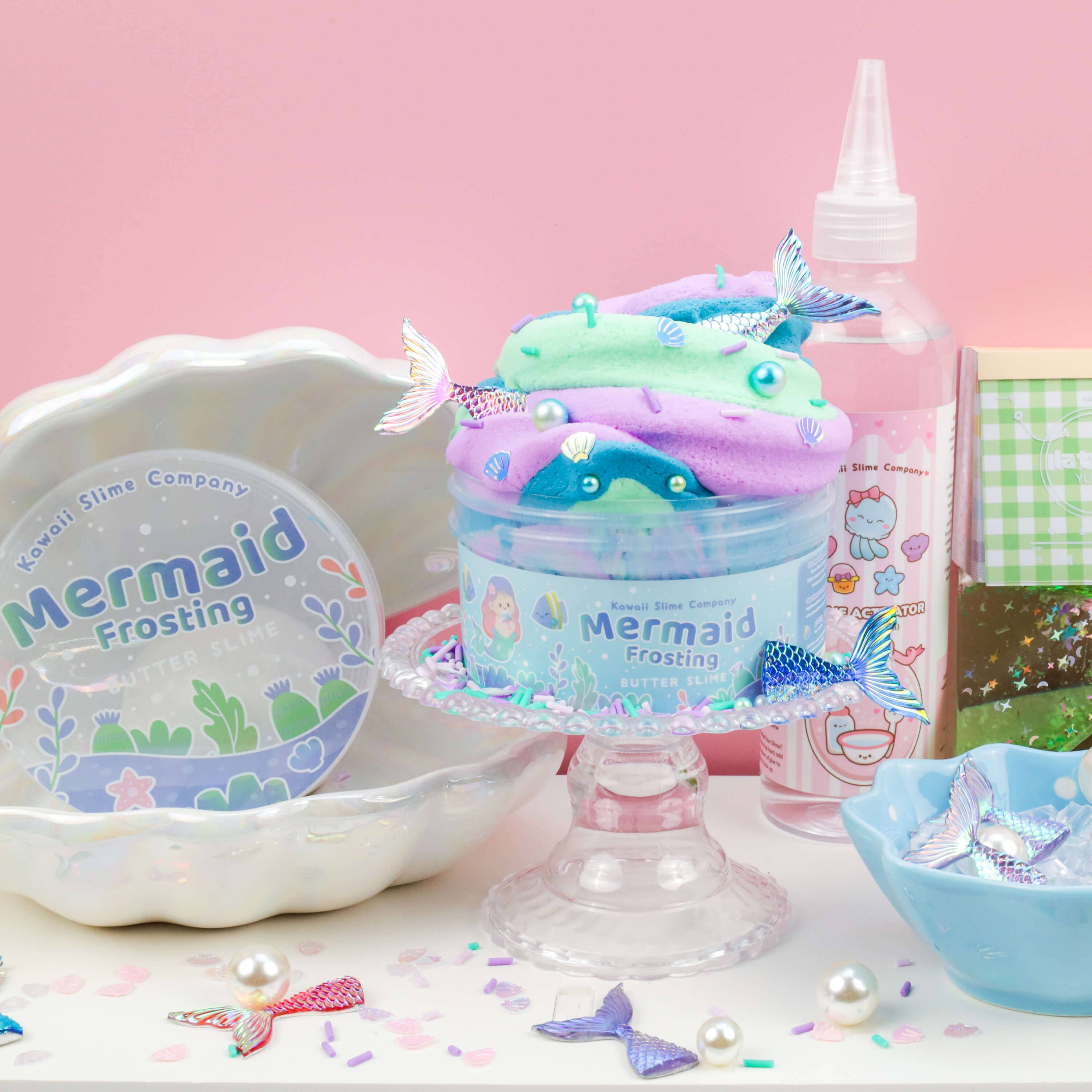 A pastel-themed setup features a jar of "Mermaid Frosting Butter Slime (4pcs/case)" by Kawaii Slime Company with a mermaid tail decoration on top, placed on a small glass cake stand. Surrounding items include a decorative dish, a spray bottle, small bowls, and more mermaid tail decorations. Confetti is sprinkled around.