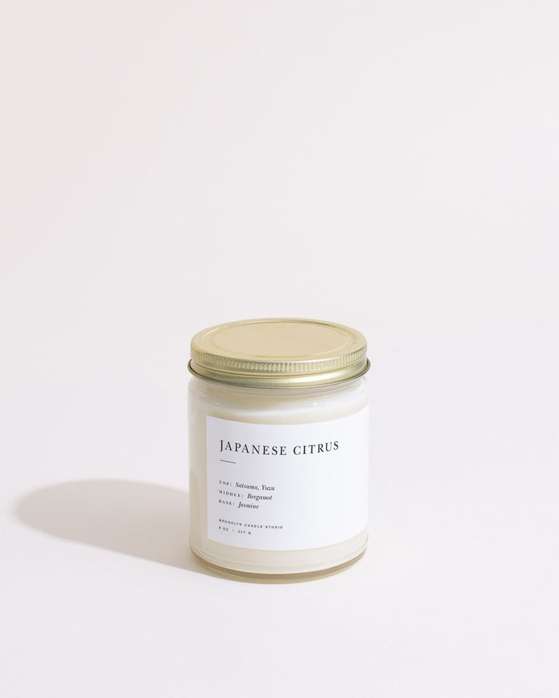 A white candle in a clear glass jar with a gold lid, featuring a minimalist label. Fragranced with satsuma, yuzu, bergamot, and jasmine. Hand-poured by Brooklyn Candle Studio for a clean burn.