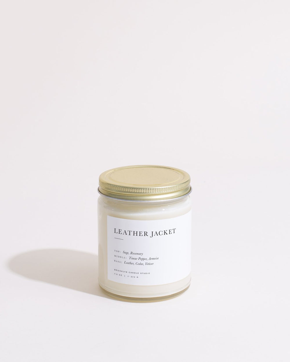 A white candle with a gold lid, part of the Leather Jacket Minimalist Candle collection by Brooklyn Candle Studio, featuring sage, timur pepper, leather, and cedar notes.