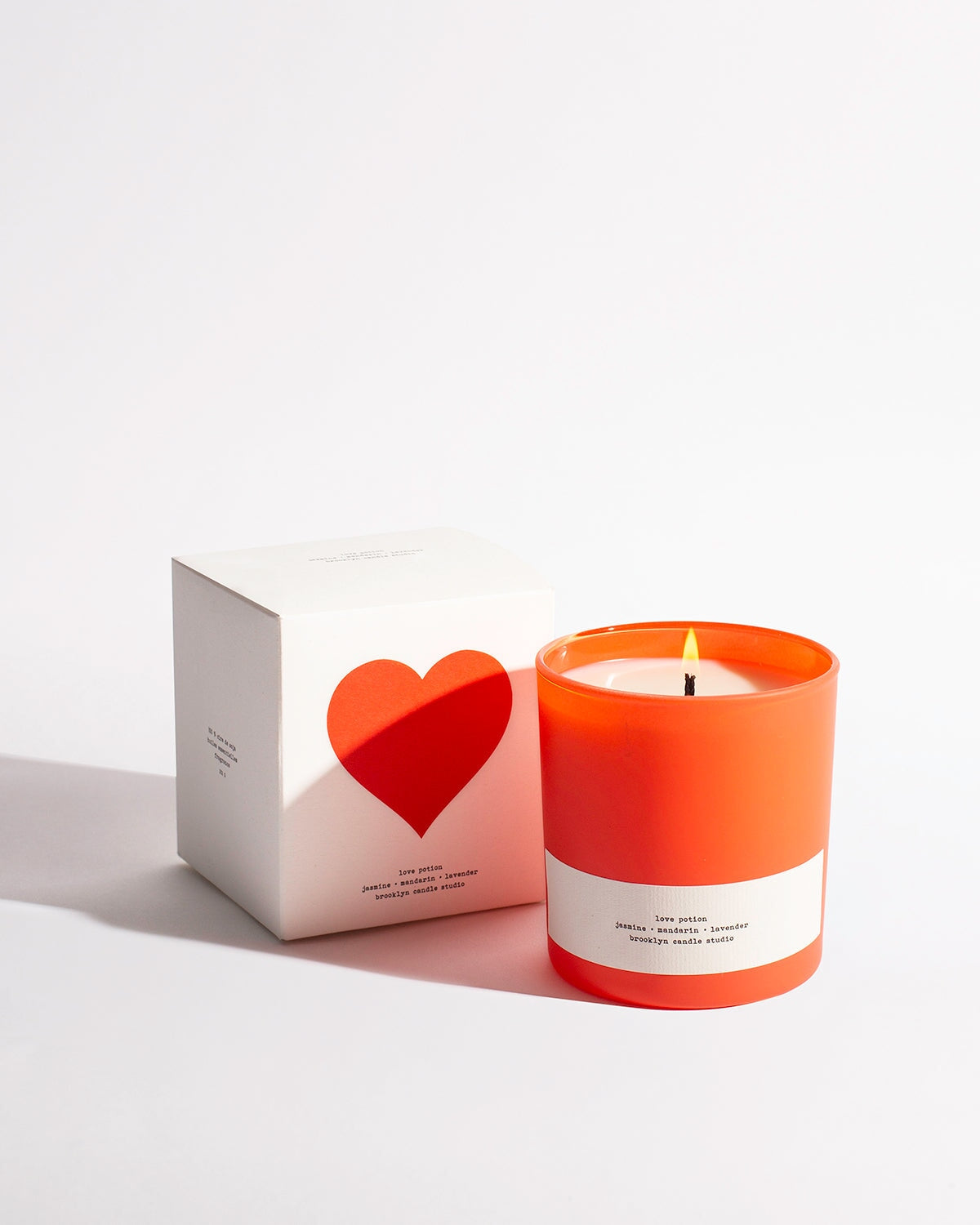 A Love Potion scented candle in a jar, nestled next to a white box with a red heart. Textured packaging inspired by vintage posters. Made in Brooklyn with soy wax.