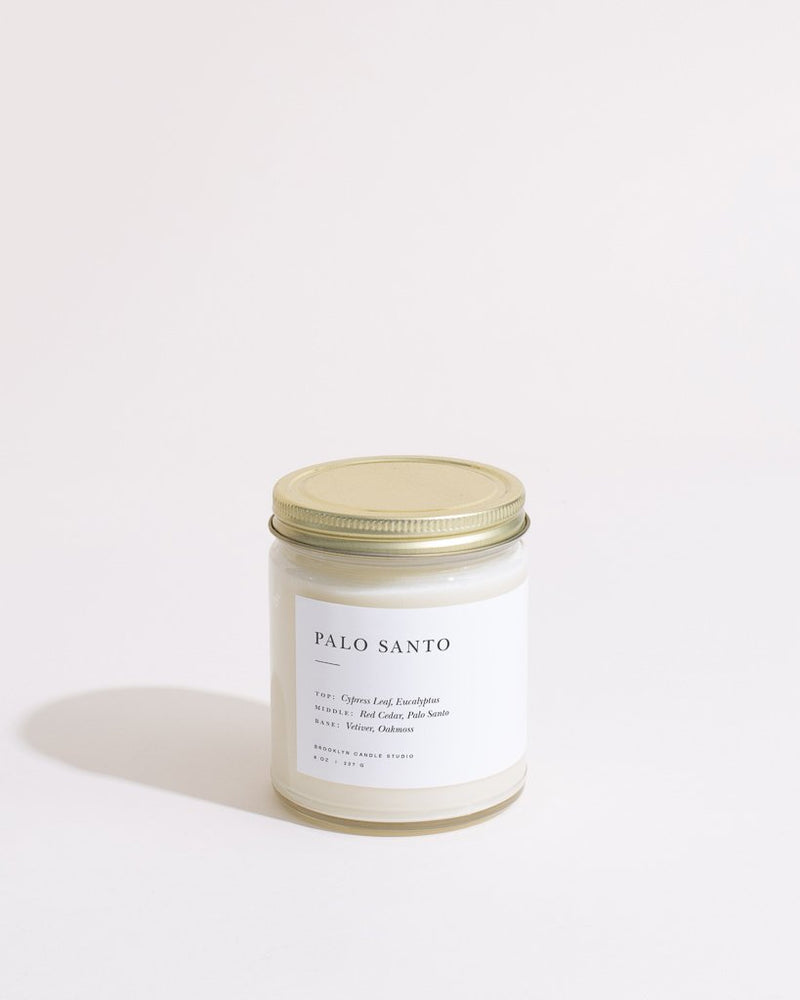 A white candle with a gold lid, featuring a label, embodies the Palo Santo Minimalist Candle by Brooklyn Candle Studio. Made with soy, cedar, eucalyptus, and vetiver notes for a 50-hour burn time.