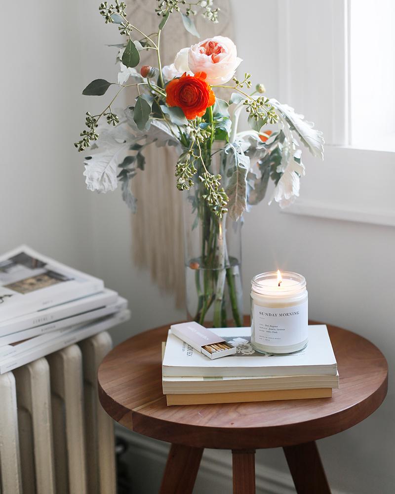 A minimalist candle in a clear glass jar, surrounded by a vase of flowers and a stack of books. Evokes a Sunday morning at the farmer's market with pear, bergamot, jasmine, gardenia, and amber woods notes.