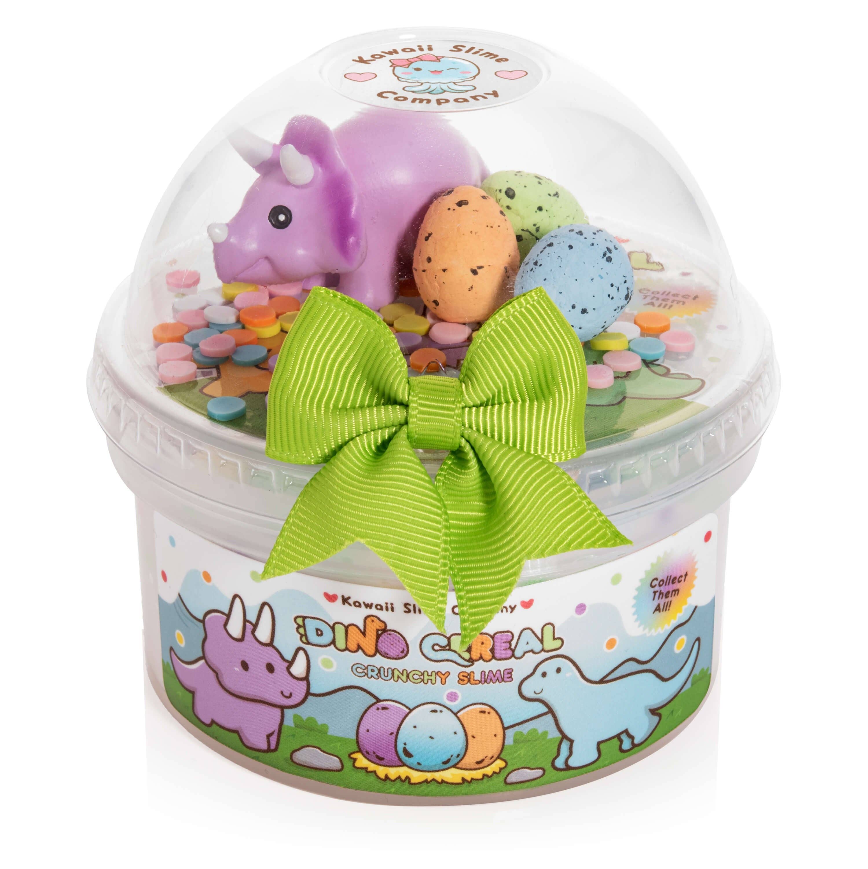 Plastic container with toy dinosaur, eggs, and green bow. Dino Cereal Crunchy Slime features glossy base, pastel foam beads, confetti sprinkles, dino eggs, and collectible dinosaur miniature.