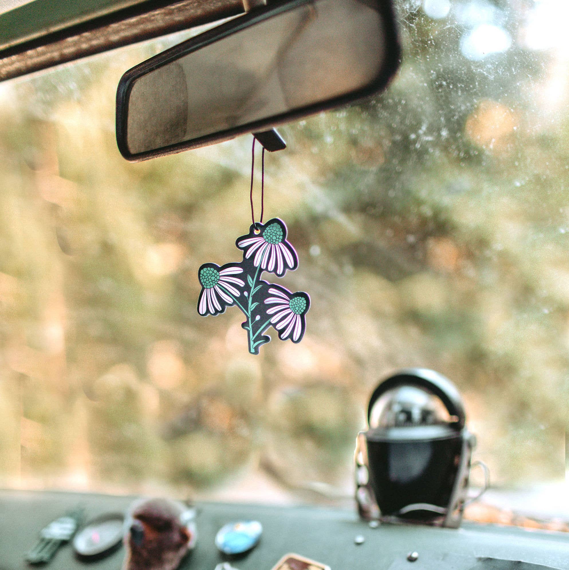 A car mirror with a Badlands Air Freshener sticker, reflecting a blurred bird's face, a black object, and a flower tag. Handcrafted auto fragrance inspired by America's national parks.