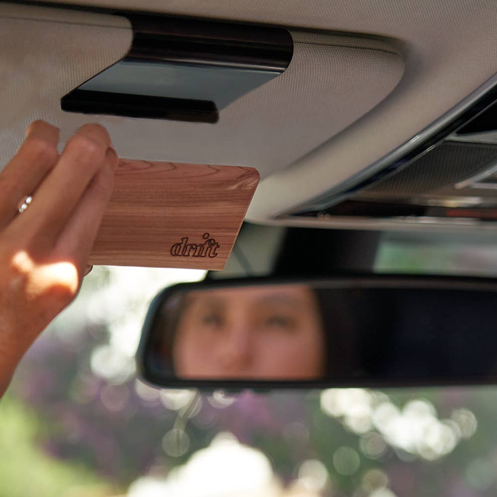 A hand holds a wooden car air freshener in a vehicle's rear-view mirror. Drift's Teak Wood Car Freshener Starter Kit includes magnetic freshener, sun visor clip, and scents like musk, amber, and cedar.