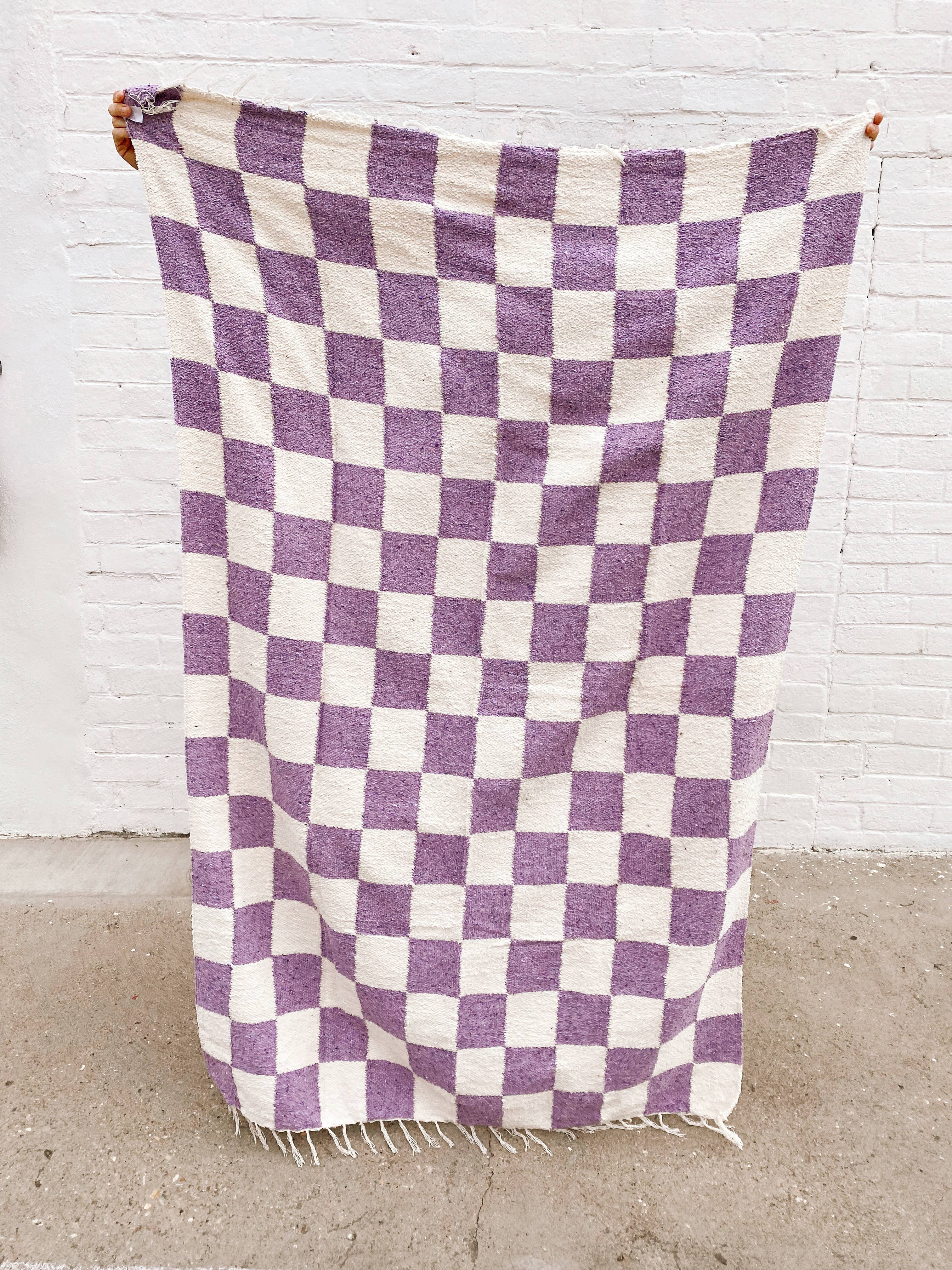 A person holding a lavender Laguna Heavyweight Checker Throw, showcasing a purple and white checkered blanket made of sustainable post-consumer materials, perfect for home or outdoor use.