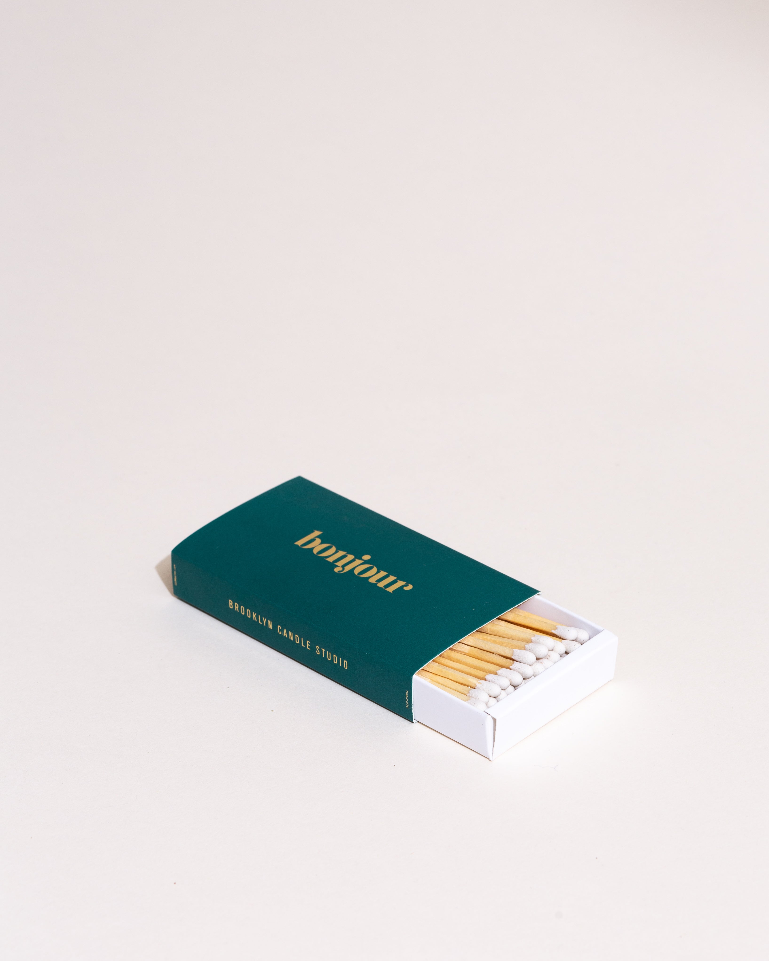 A box of XL Statement Matches - Bonjour with gold foil-pressed cheeky greetings, containing 55 white-headed matches, on a white background. Dimensions: 4.7” x 2.5” x 0.78”.