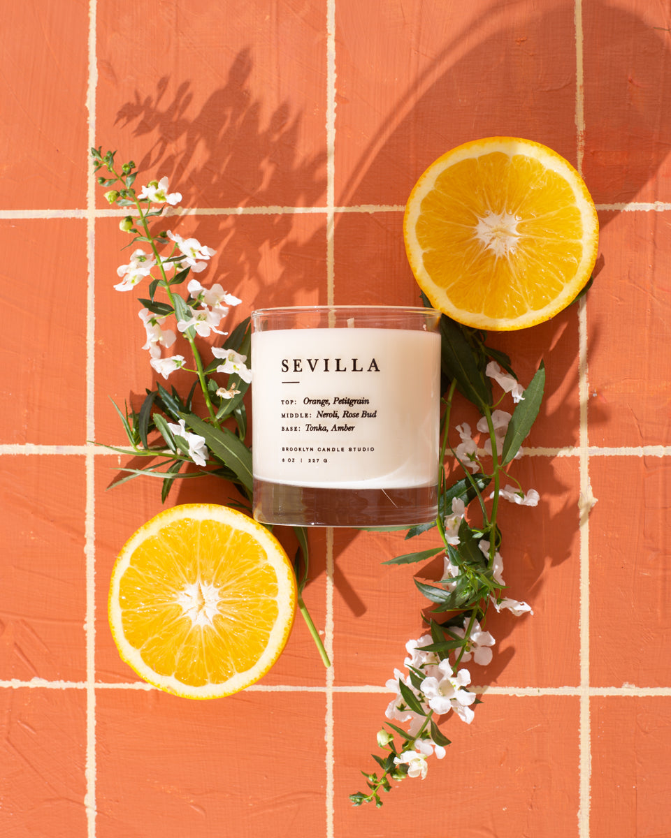 A close-up of the Sevilla Candle featuring orange slices, flowers, and a label on a tile surface, embodying citrus and incense notes for a captivating, eco-friendly soy wax experience.