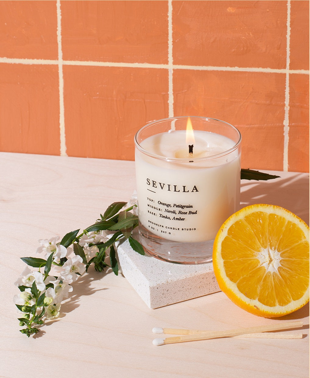 A candle in a glass jar surrounded by citrus fruits on a wooden surface, embodying Sevilla's blend of orange blossoms, incense, and flamenco-inspired vibes from Brooklyn, New York.