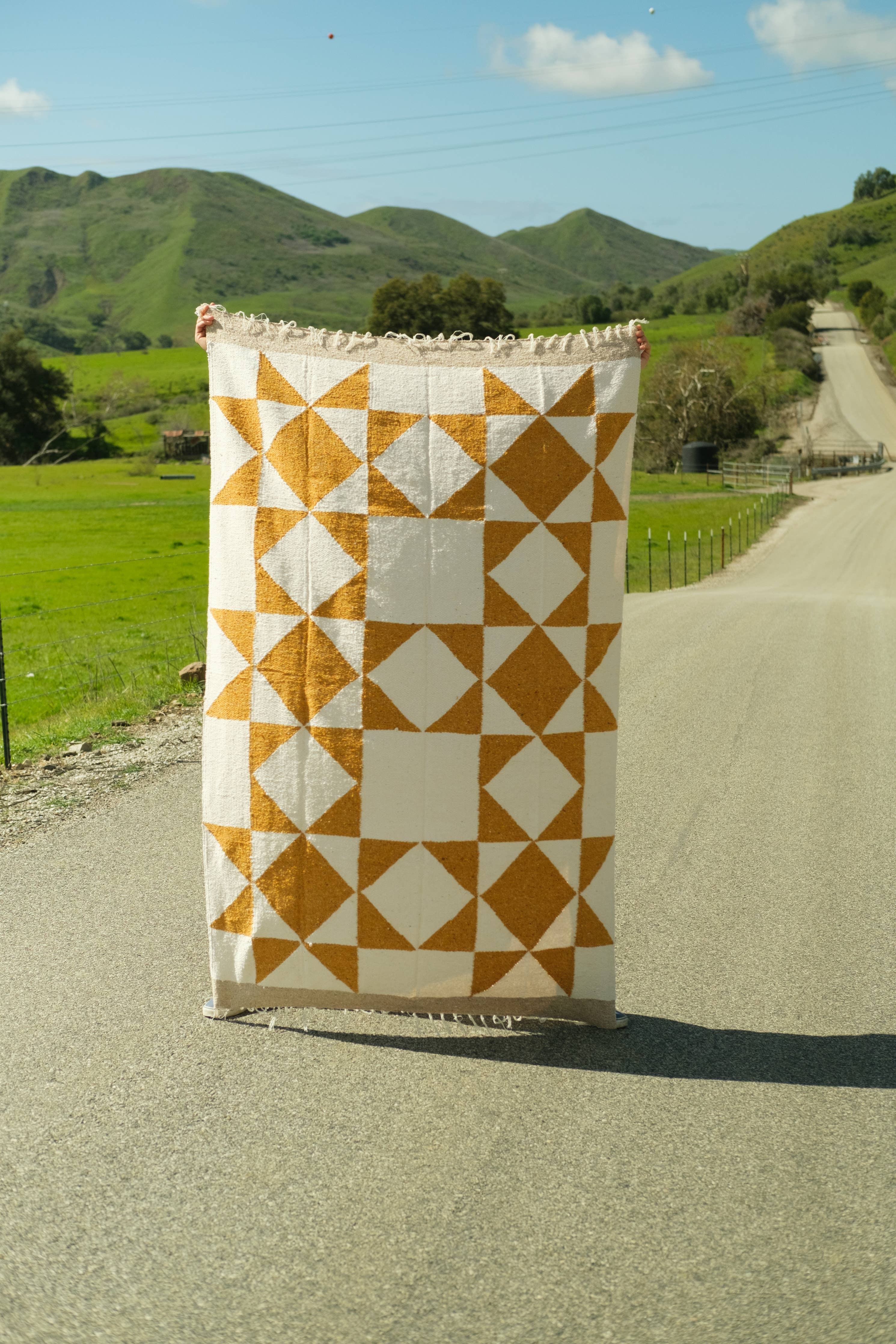 A person holds a Gold & Cream Patchwork Sun blanket on a road. Handmade from reclaimed fibers, this heavyweight throw measures 83 x 52 inches. Designed in California, artisan made in Mexico.