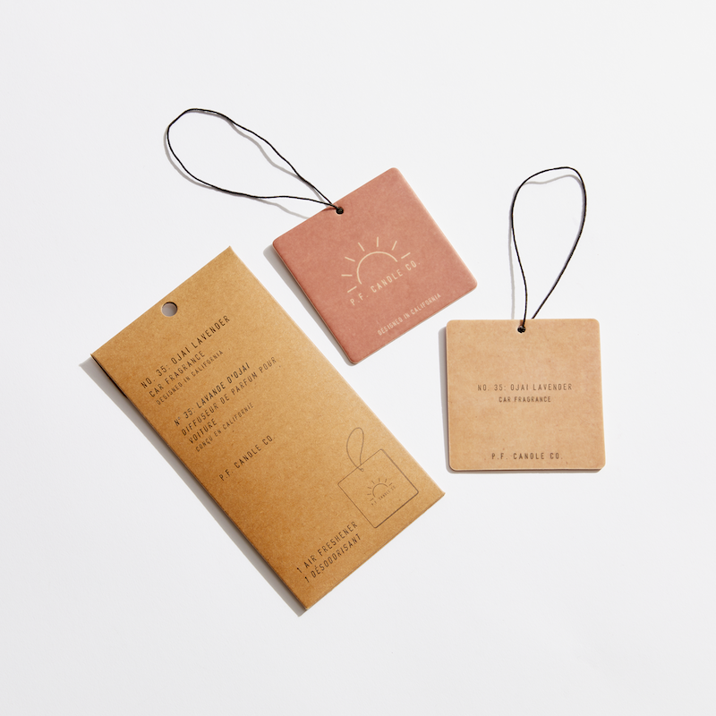 A group of Ojai Lavender car fragrance tags with strings, featuring a brown paper tag, close-up of a logo, and pink tag with black string. Sold wholesale at $4.50/unit.