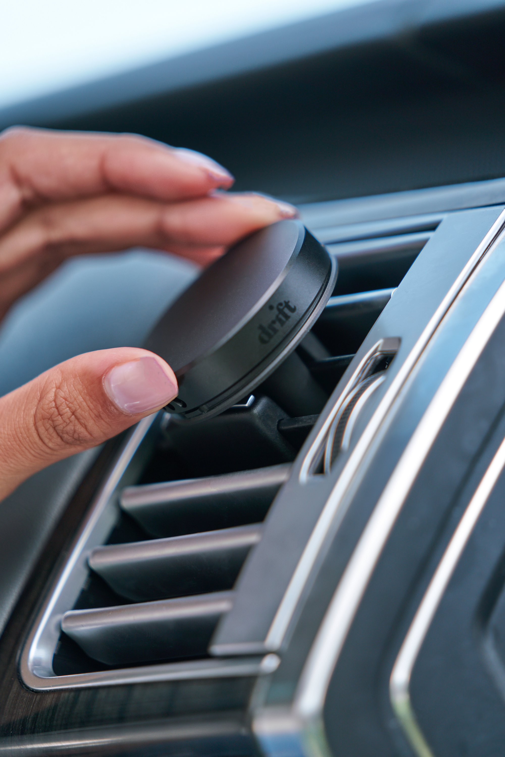 A hand holds Metal Car Vent Freshener - Teak, a black alloy air freshener blending with a car's interior. Includes 3 scent refills.