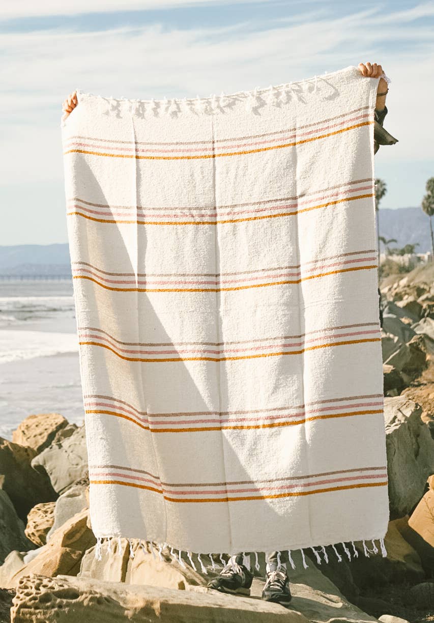 Person holding a sustainable Solimar throw blanket outdoors, featuring natural base color with mustard, pink, and tan stripes. Made from reclaimed fibers for durability and softness. Measures 73 x 48 inches.